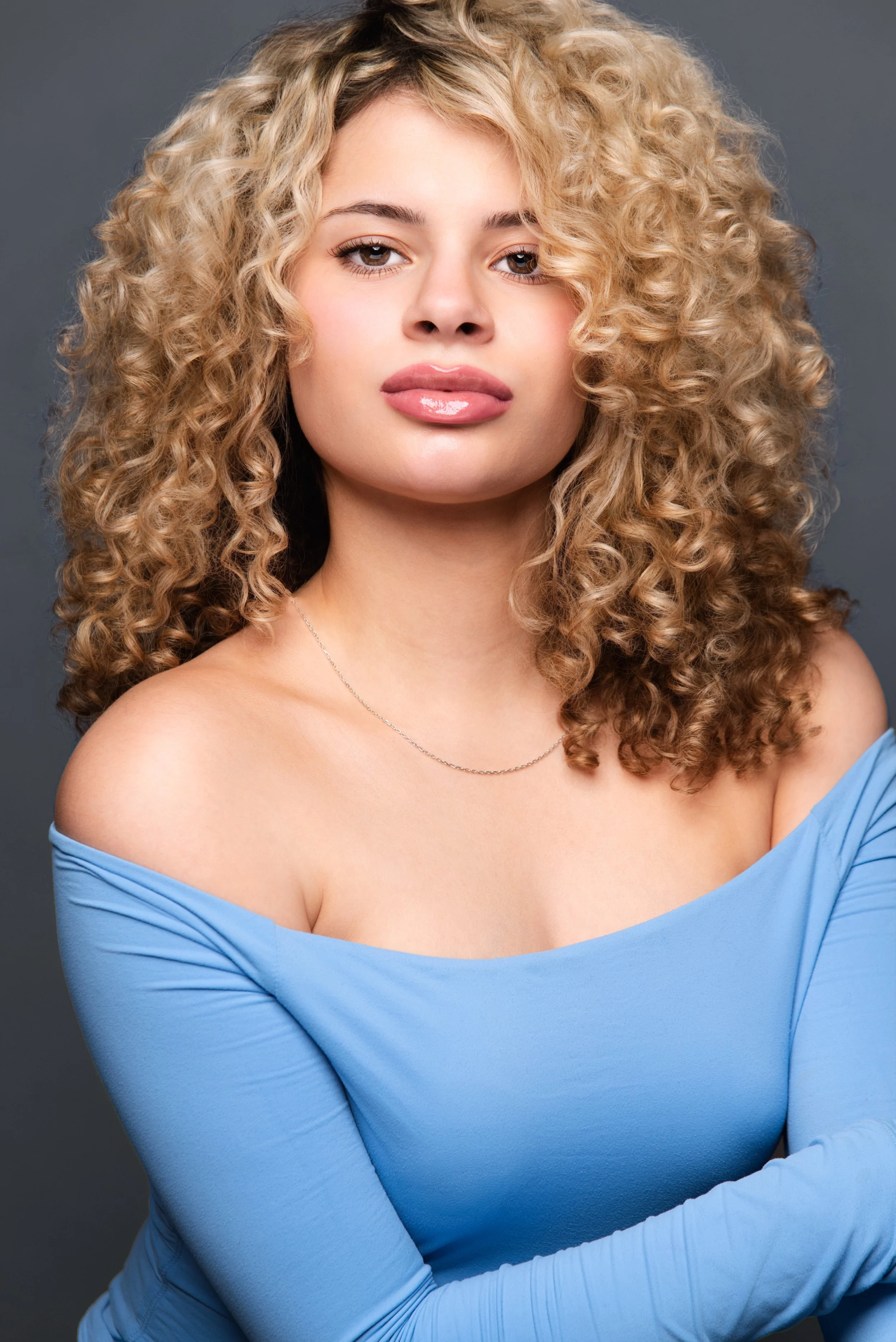 A young woman with curly blonde hair and light skin wearing a light blue off-the-shoulder top and a silver necklace, posing against a gray background. Darius Nichols Photography DNPhotographyNYC