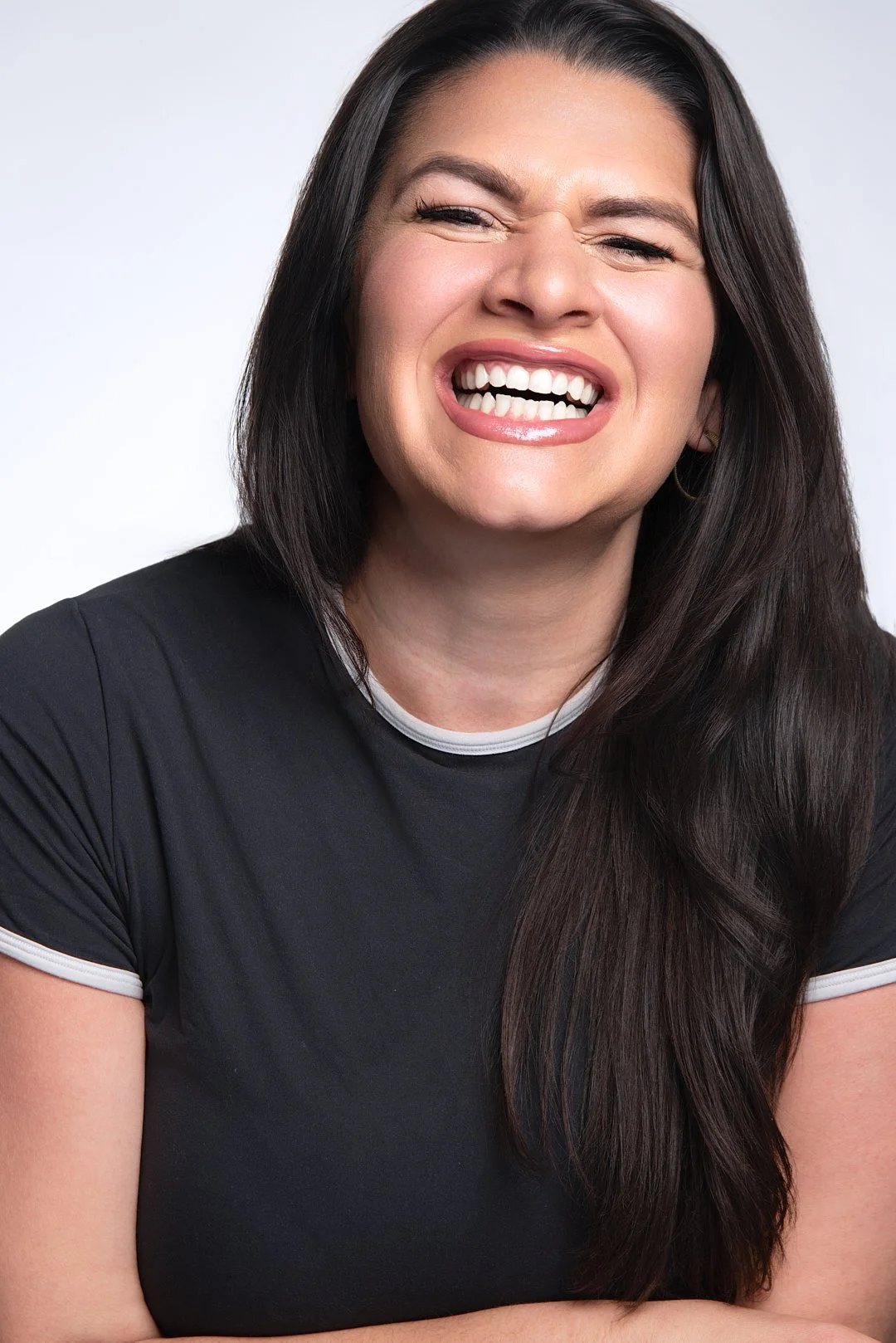 A woman with long dark hair smiling and showing her teeth, wearing a black shirt with white trim on the collar and sleeves, against a plain white background.