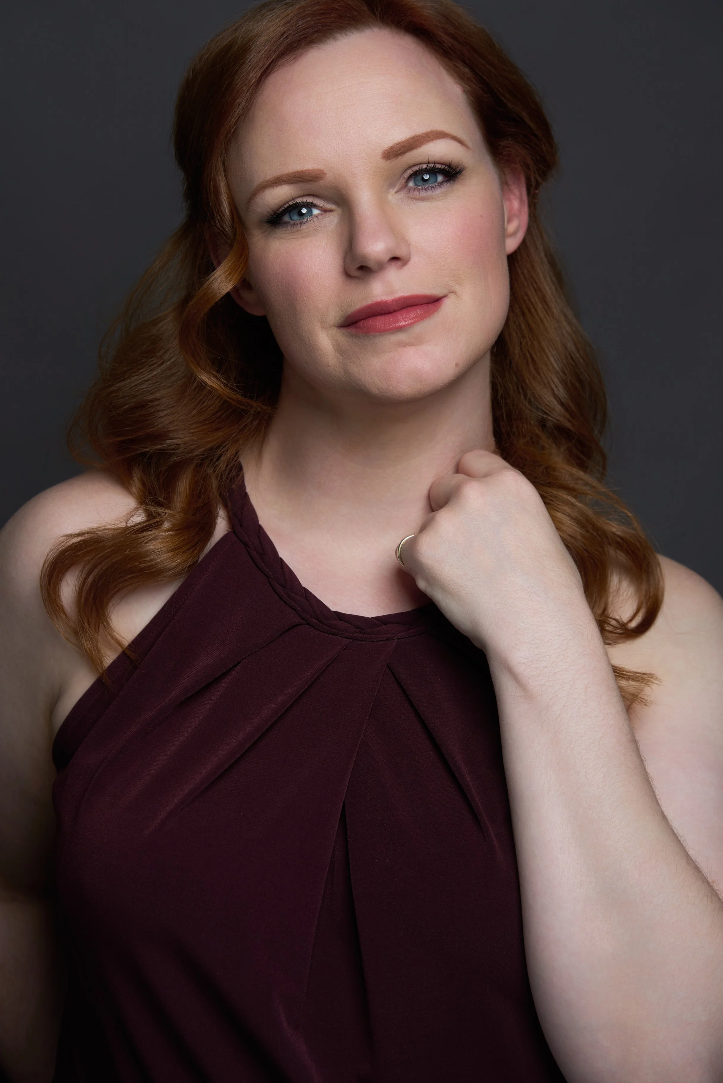 A woman with red hair, blue eyes, and wearing a burgundy sleeveless dress, posing against a dark background.