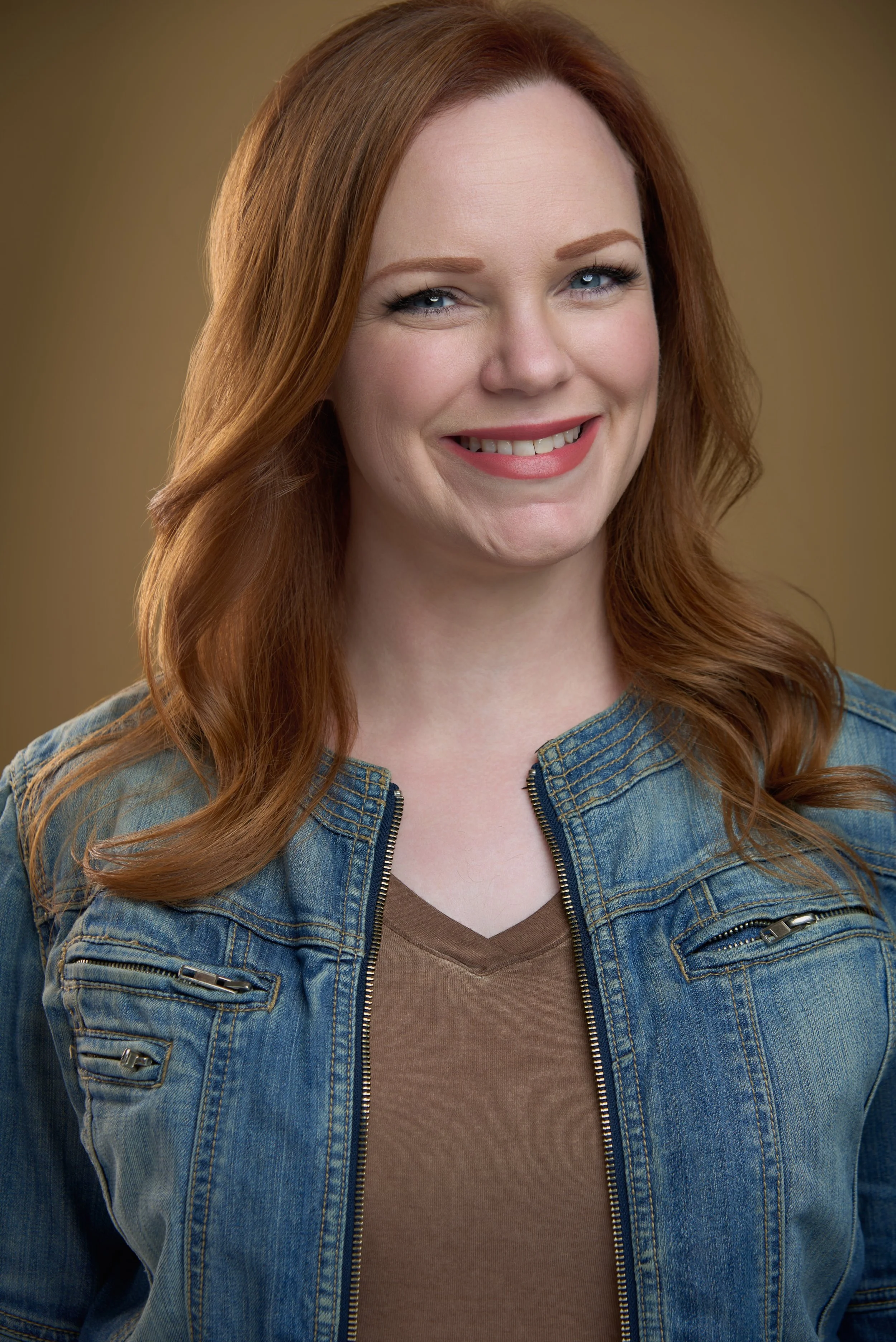 A woman with long red hair, blue eyes, wearing a denim jacket over a brown top, smiling at the camera.