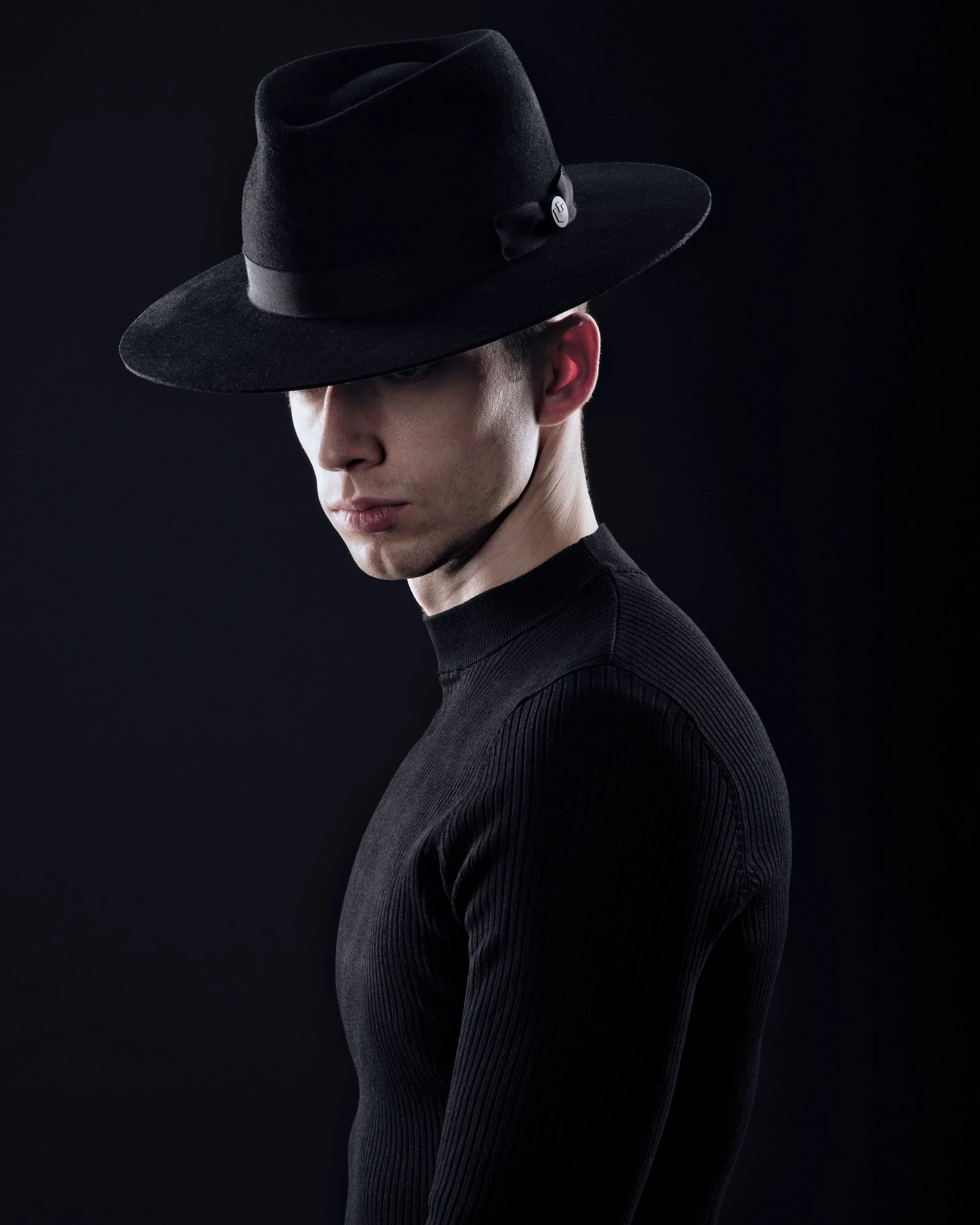 A young man wearing a black wide-brimmed hat and a black ribbed turtleneck against a black background, looking downward.