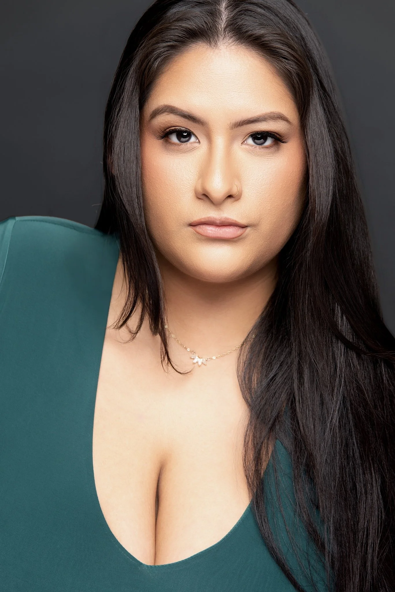 A woman with long dark hair, wearing a deep neckline green top, and a delicate necklace, looking directly at the camera against a dark background. Darius Nichols Photography DNPhotographyNYC