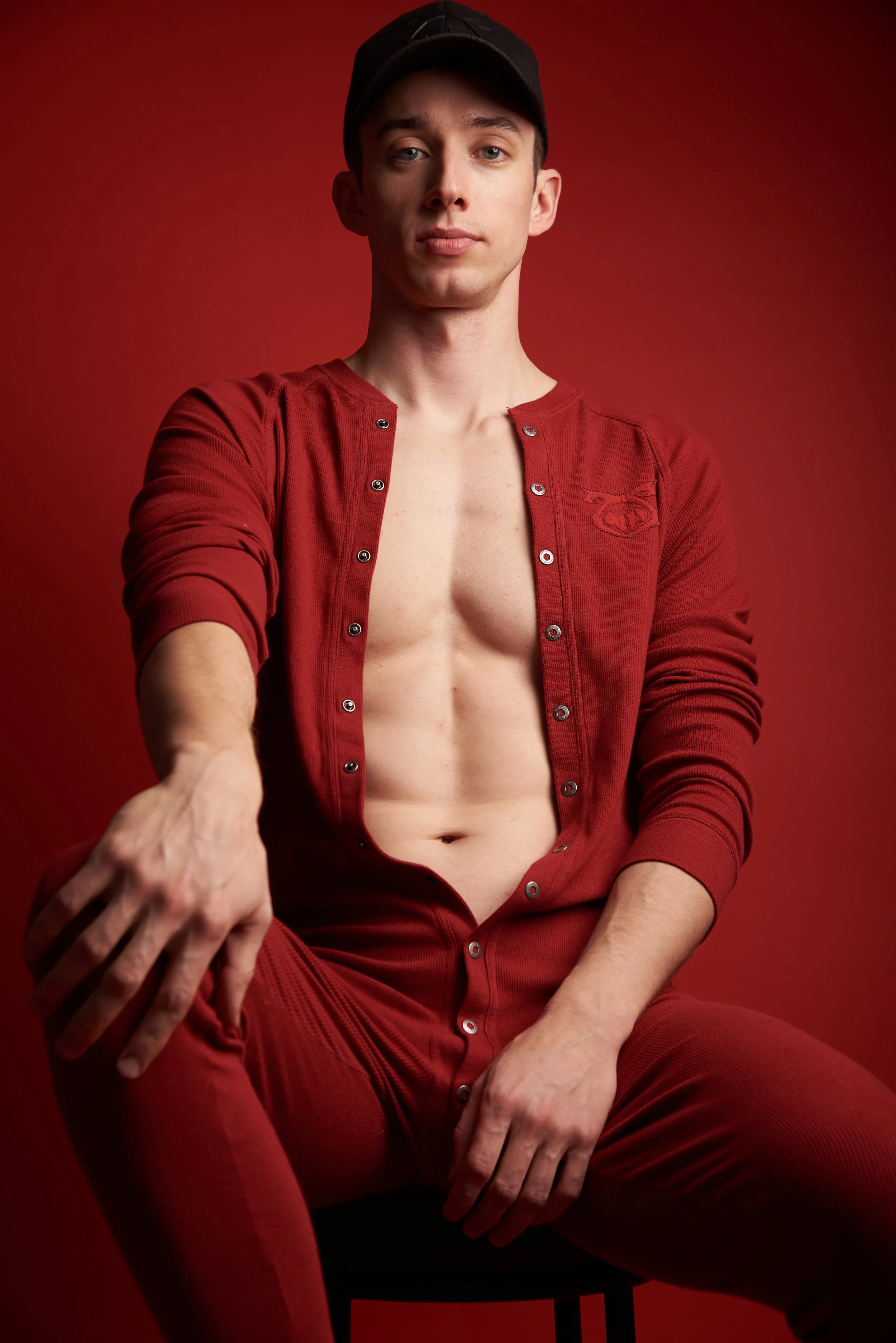 A young man wearing a black cap and a red button-up shirt with the buttons undone, revealing his torso, sitting against a matching red background.