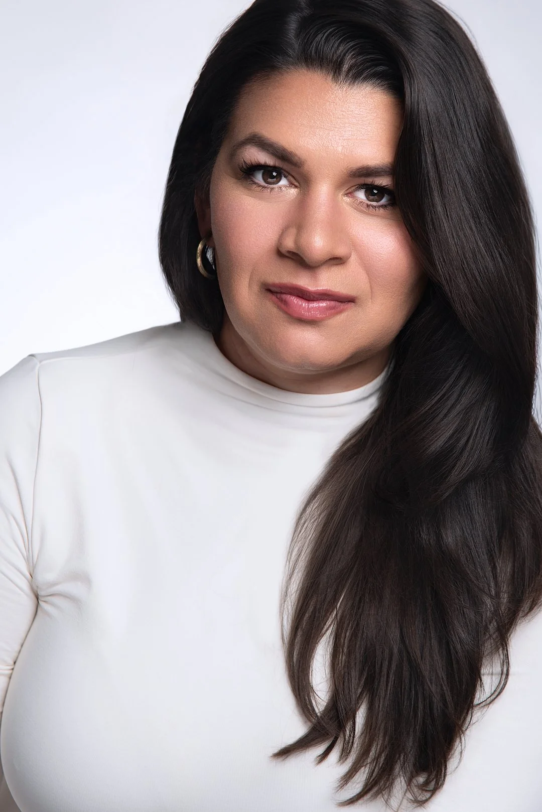 Close-up portrait of a woman with long dark hair, wearing hoop earrings and a white top, looking confidently at the camera against a light gray background.