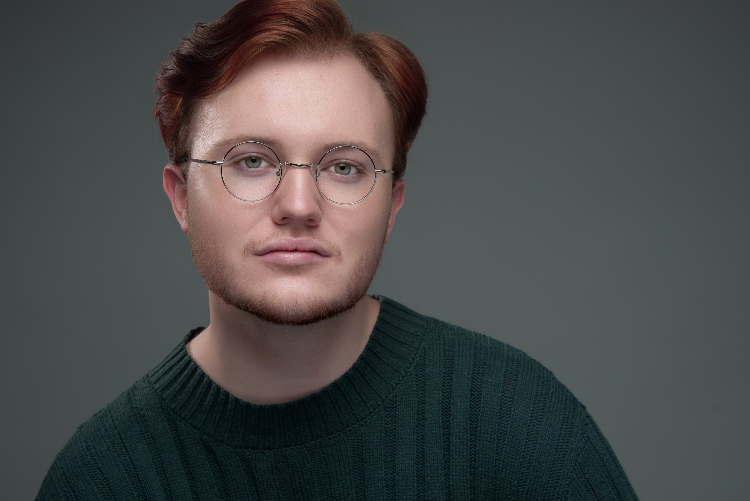 Portrait of a young man with red hair, glasses, and a light beard wearing a dark green sweater against a gray background.