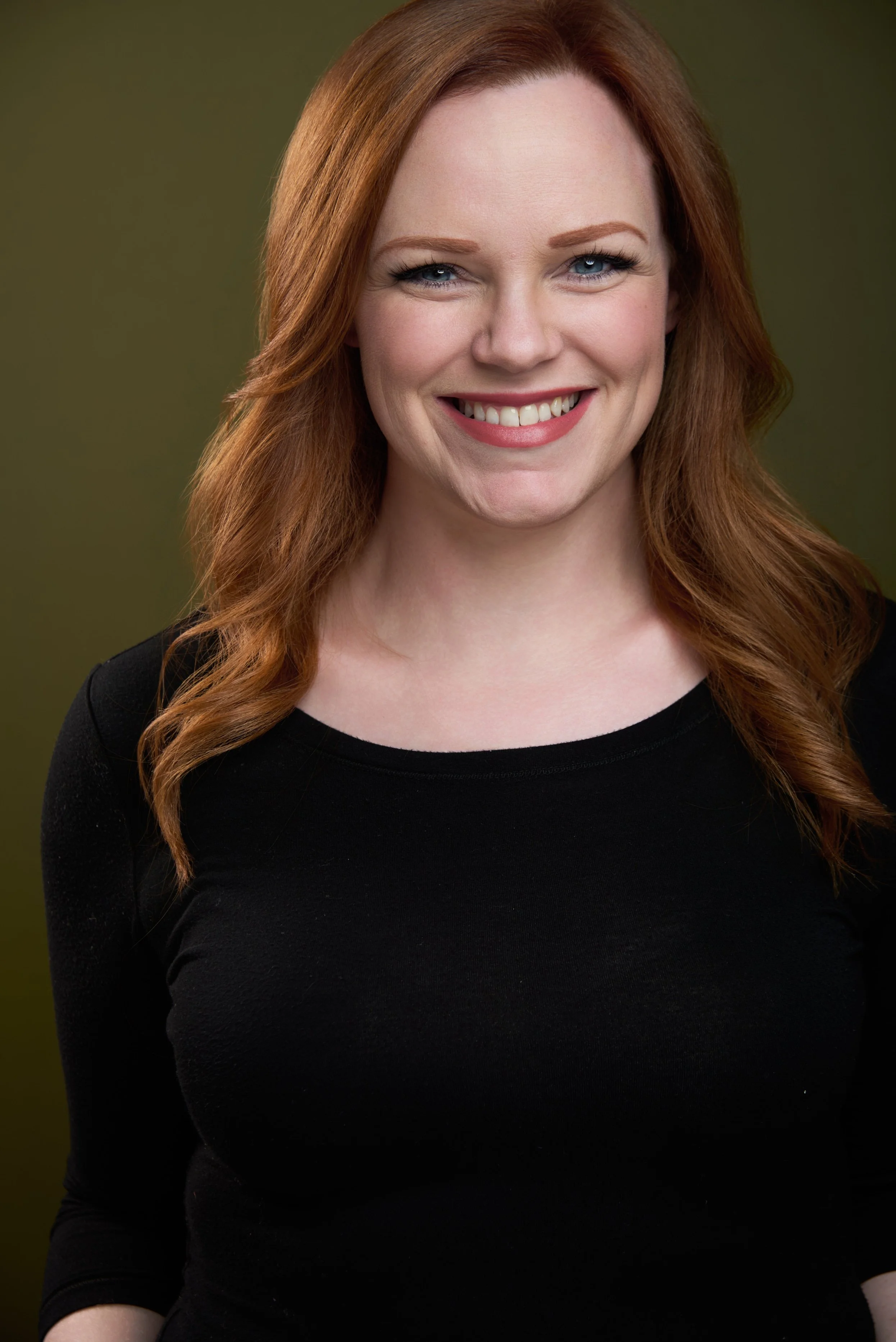 A woman with red hair, blue eyes, and fair skin, smiling, wearing a black top, with an olive green background. Darius Nichols Photography DNPhotographyNYC