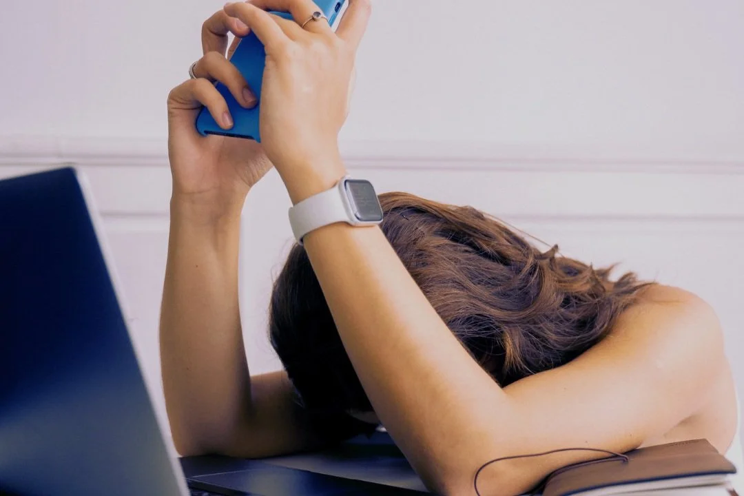 A woman with brown hair is resting her head on her arms on a desk, holding a smartphone above her head. She is wearing a smartwatch.