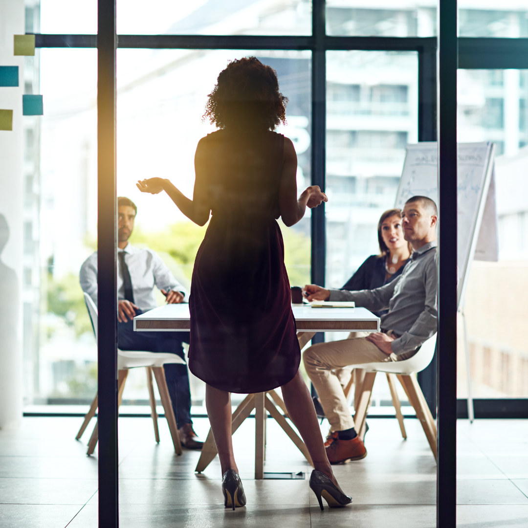 A woman in a black dress and high heels stands in front of a glass conference room with three seated colleagues, two men and one woman, during a meeting or presentation.