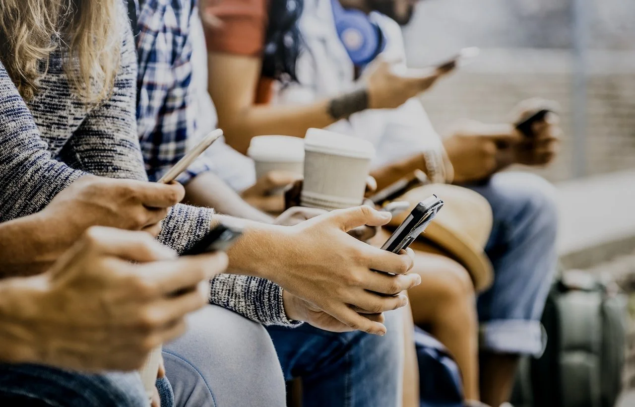 Group of people sitting outdoors, each holding their smartphones and some with coffee cups.