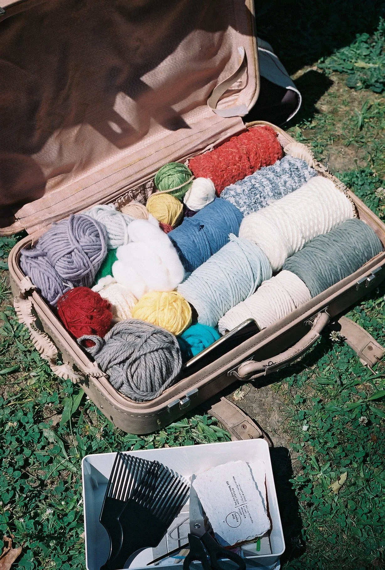 Open vintage suitcase filled with colorful balls of yarn and knitting supplies on grass, with a pair of scissors and comb in a white container nearby.
