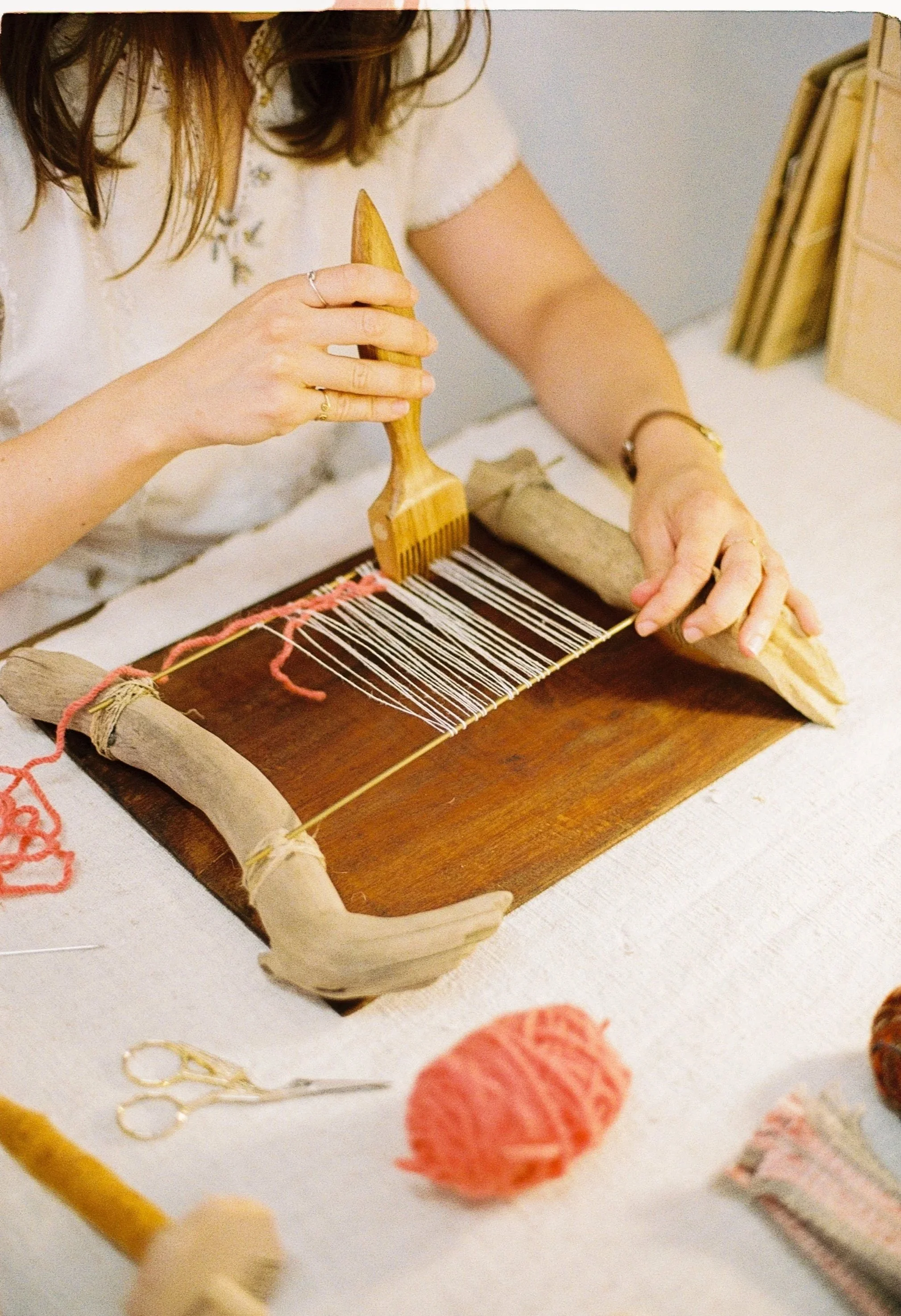 Maria Elisa Vale weaving on a small loom, surrounded by balls of yarn and weaving supplies on a table.
