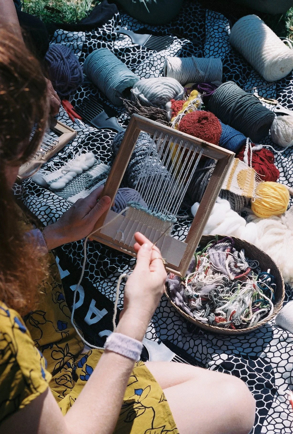 Person weaving yarn on a wooden frame, surrounded by various skeins of yarn and knitting supplies on a patterned blanket.