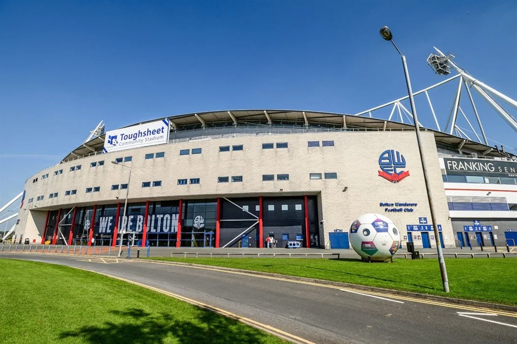 Exterior of a large sports stadium with the words "WE HOPE BOLTON" displayed on windows, a sign for Toughsheet Community Stadium, and a logo for Bolton Wanderers Football Club. There is a large white sphere sculpture and a grass area in the foreground.