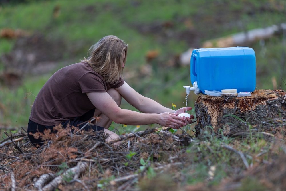 Person washing their hands using water trickling from a portable water tank propped on a tree stump.