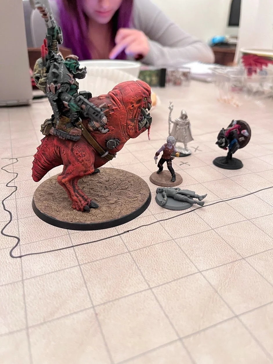  Three miniature figurines stand in battle poses against another figurine, of a giant worm with legs and teeth. A fifth figurine lays on their back, seemingly unconsious. 