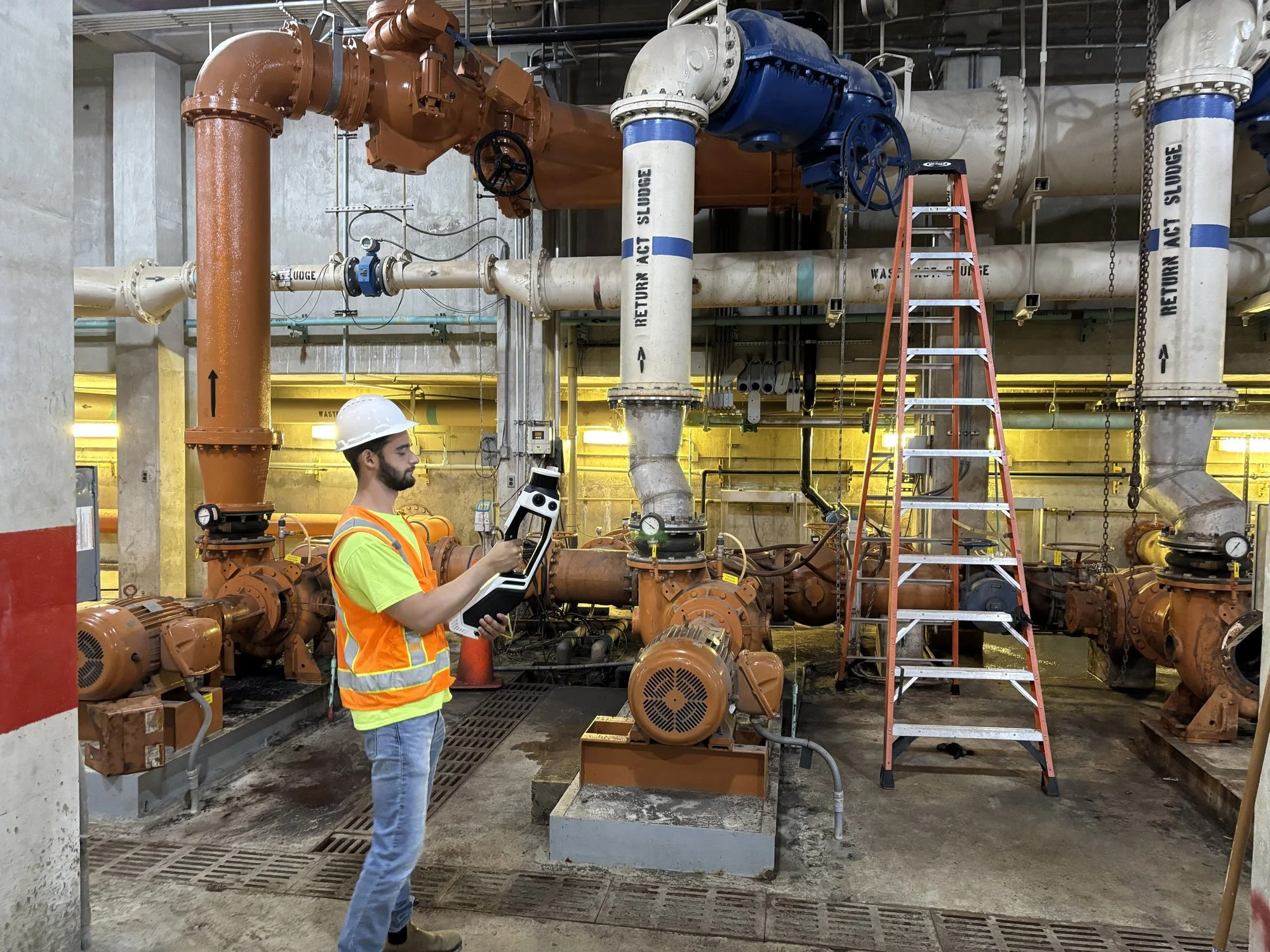 A man in a hard hat and safety vest stands in an industrial plant with large orange and white pipes, valves, and machinery. He is holding a tablet or device, and there is a ladder nearby.