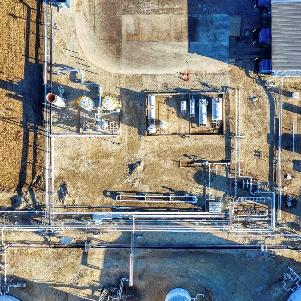 An aerial view of an industrial facility with pipelines, storage tanks, and equipment on a dirt ground, with a building in the upper right corner and shadows cast by the structures.