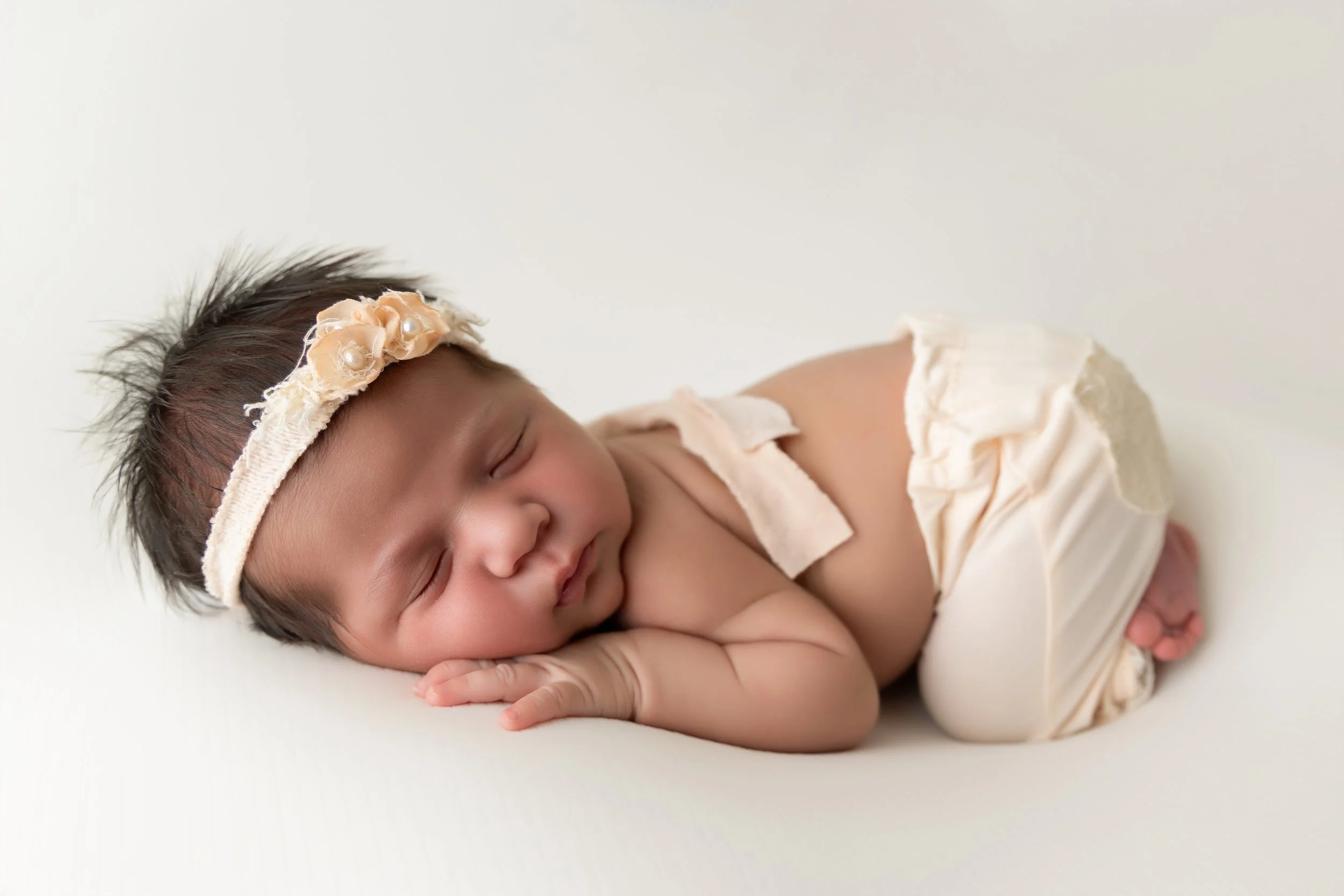 Newborn baby wearing white knit overalls laying on a white plush with a plant next to him