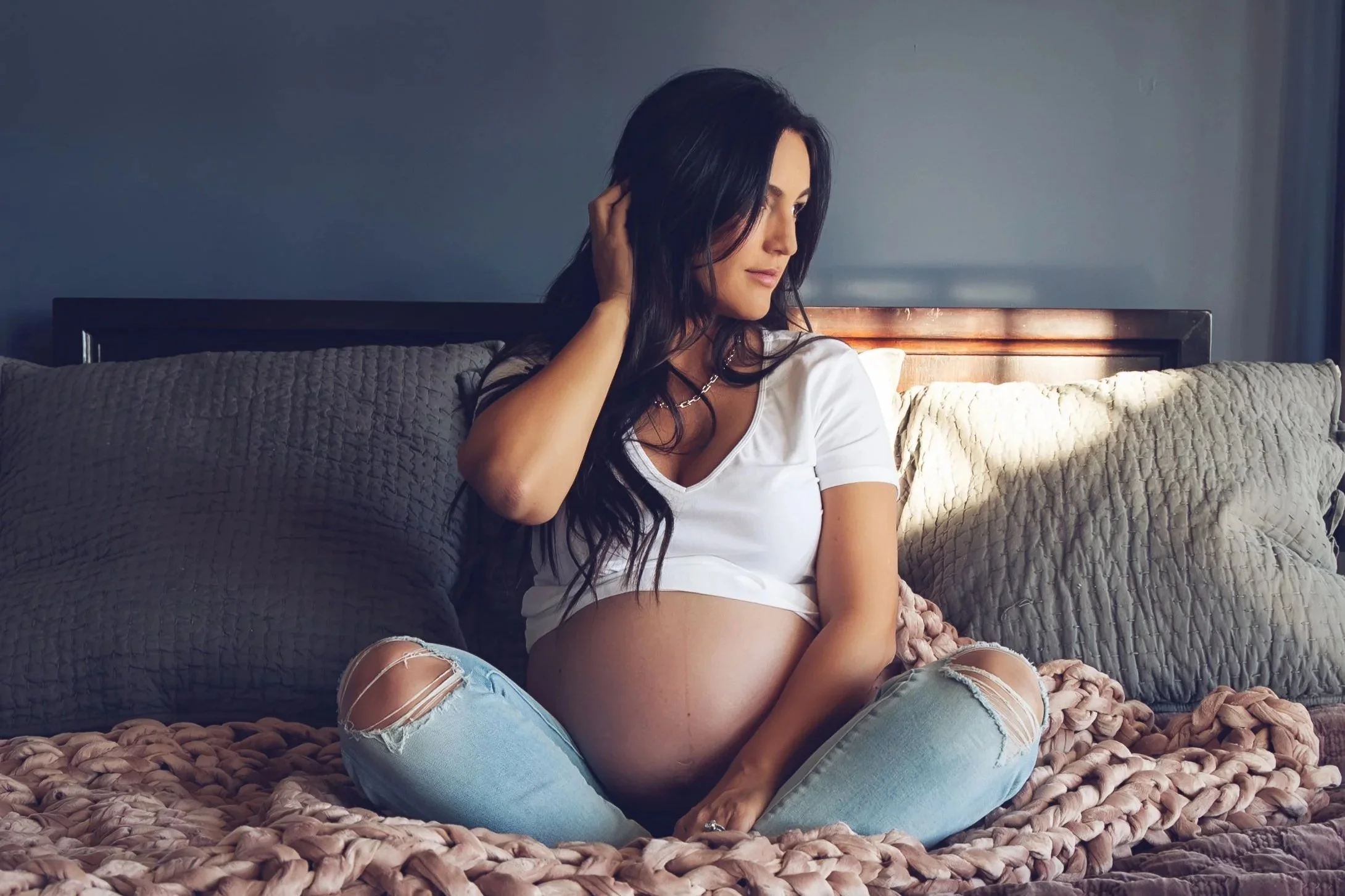 Woman sitting on a bed with a long dark brown hair with a bare pregnant belly showing underneath a white crop top