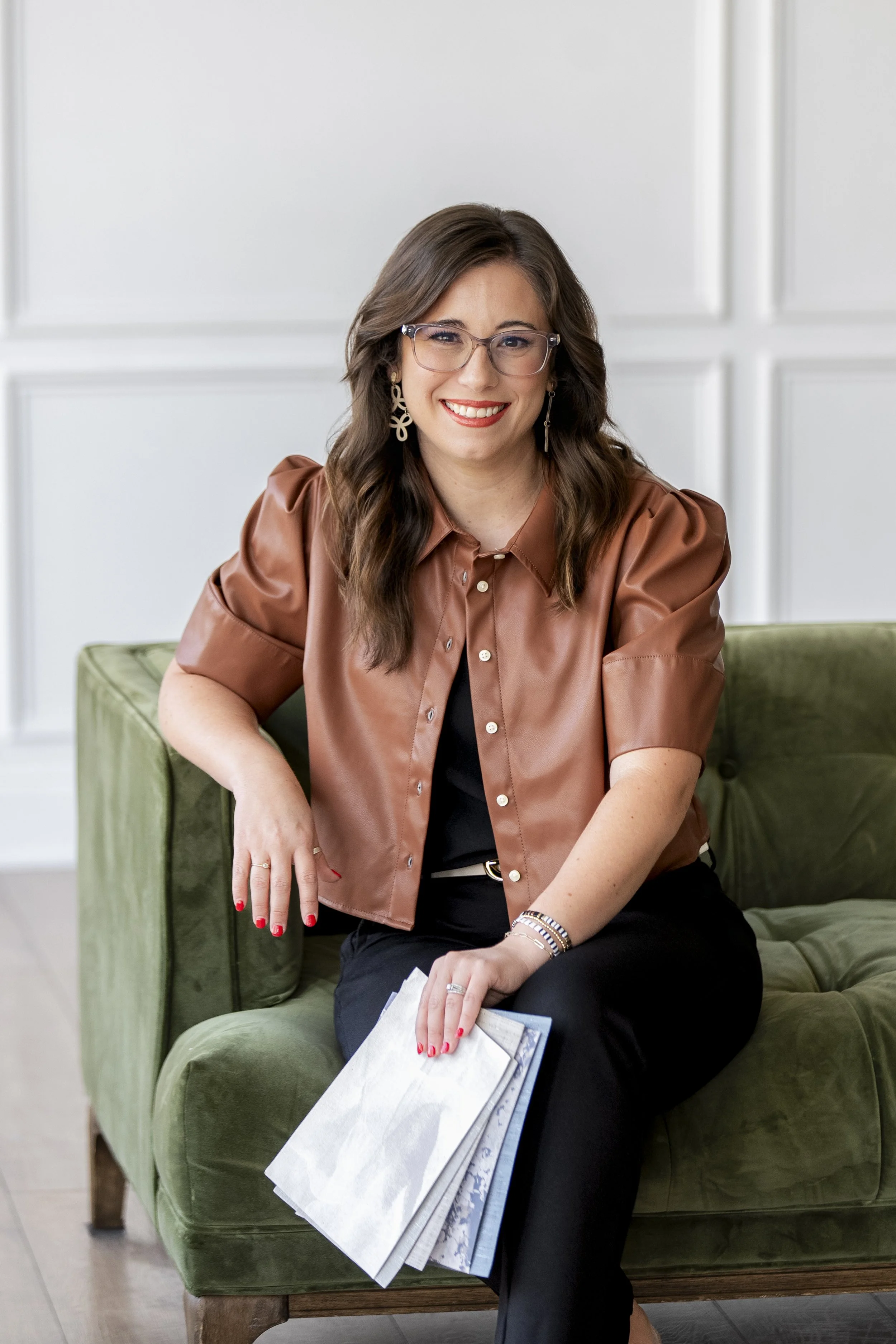 A woman with brown hair, glasses, and earrings sitting on a green couch, smiling, holding papers with her left hand resting on her knee.