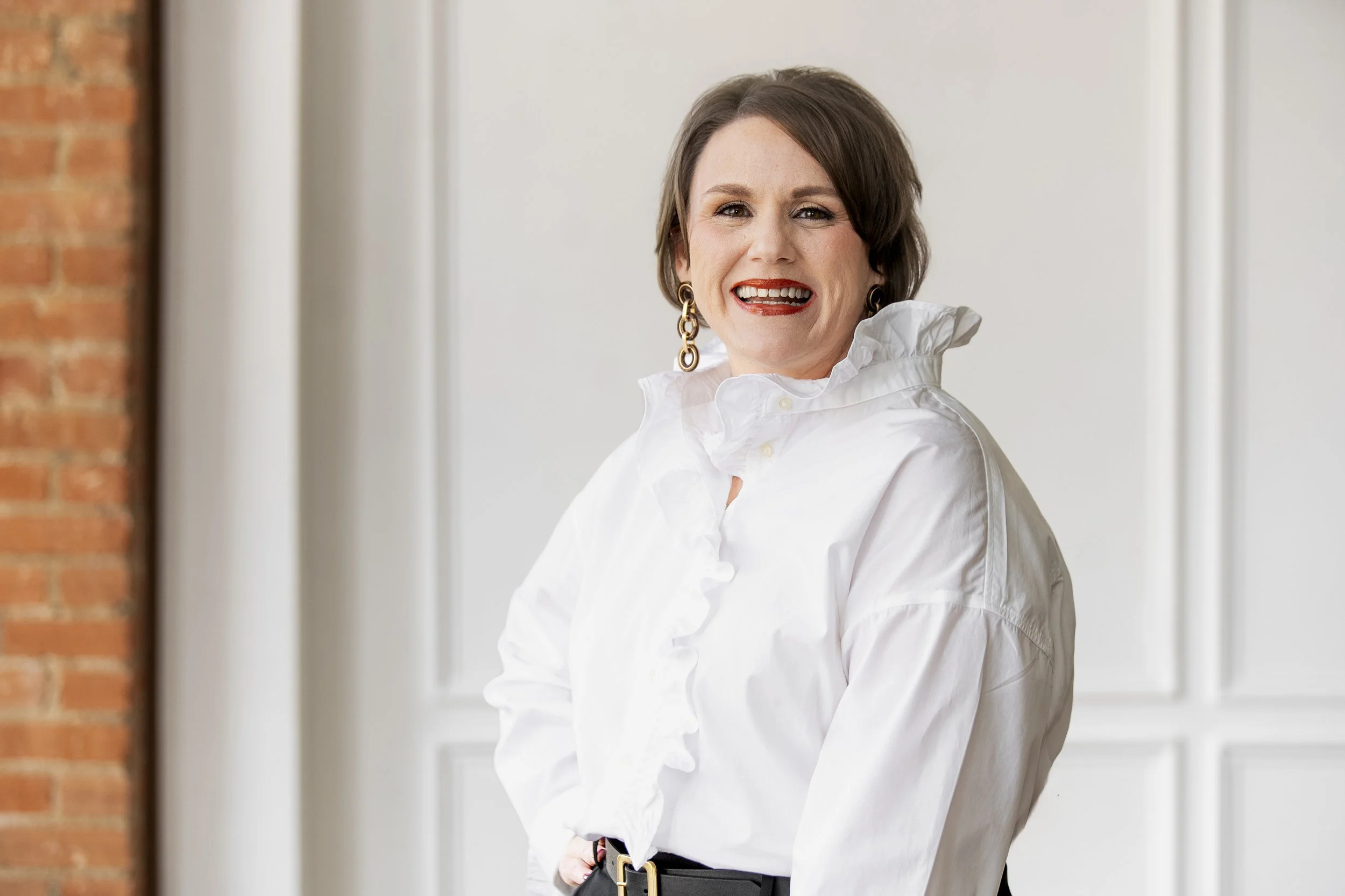 A woman with short brown hair and earrings smiling in a white ruffled blouse, standing in front of a brick wall and white paneled wall.