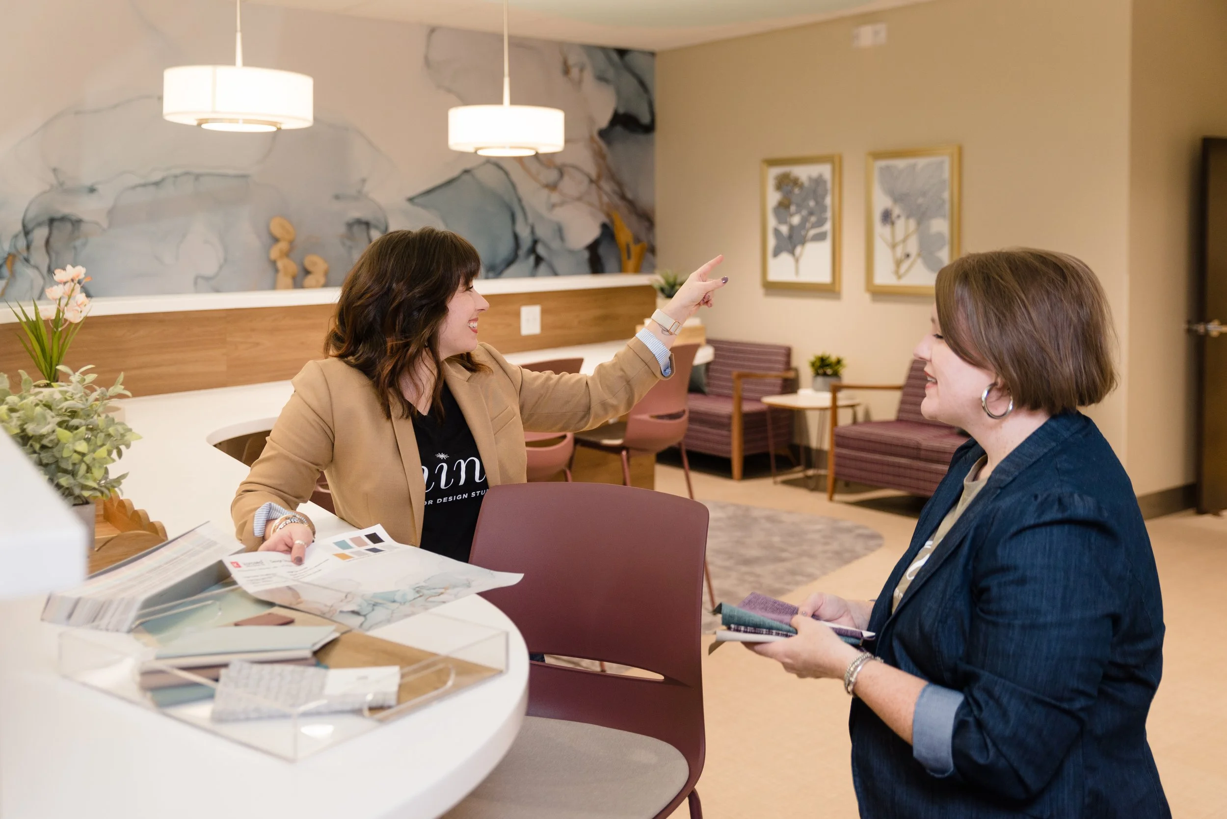 Two women sitting at a table engaged in conversation in a modern, well-lit room with artwork and plants, one woman pointing and the other holding a magazine.