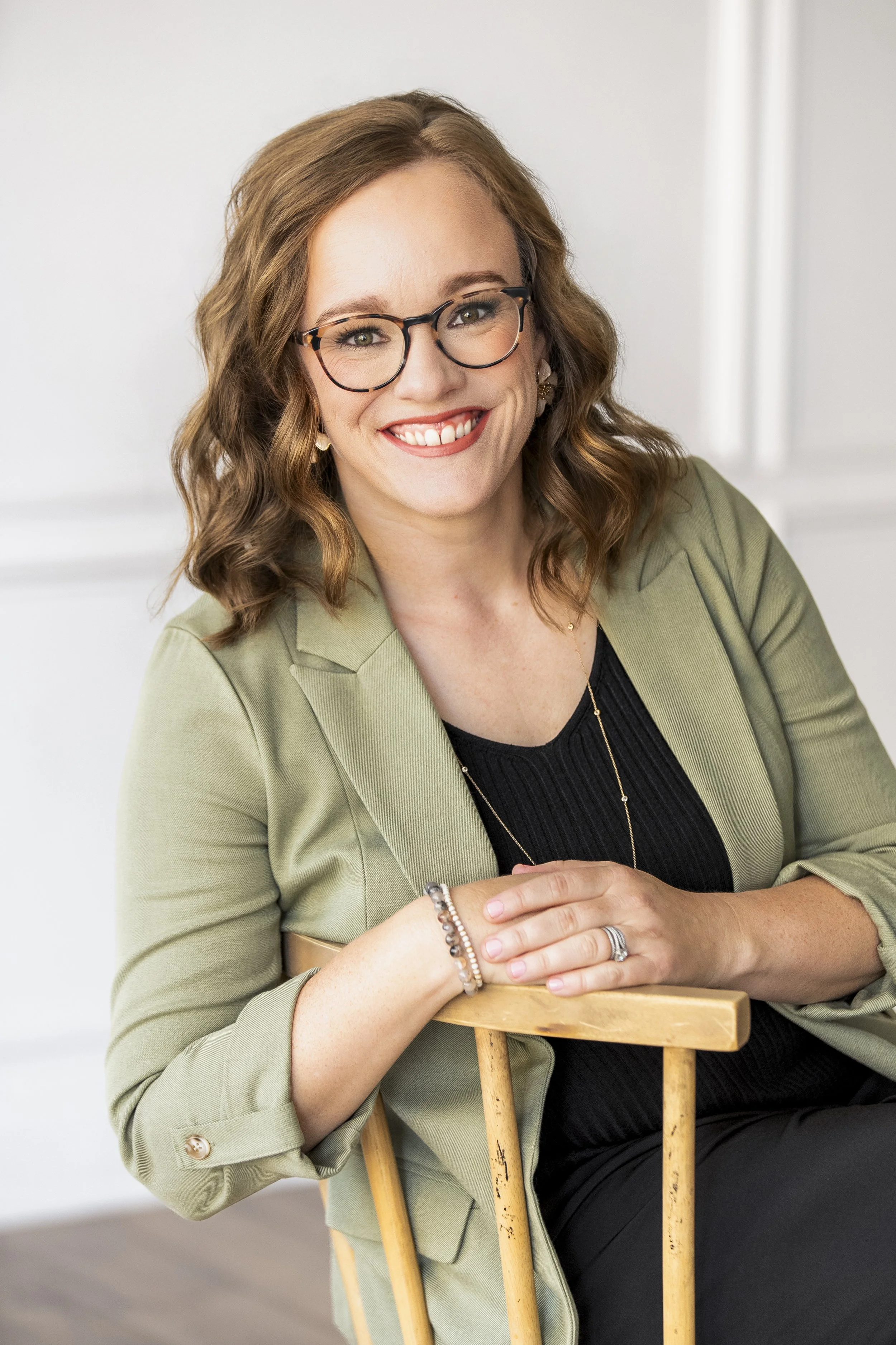 A woman with shoulder-length wavy red hair, wearing glasses, a black top, and a beige cardigan, smiling and sitting at a wooden table.