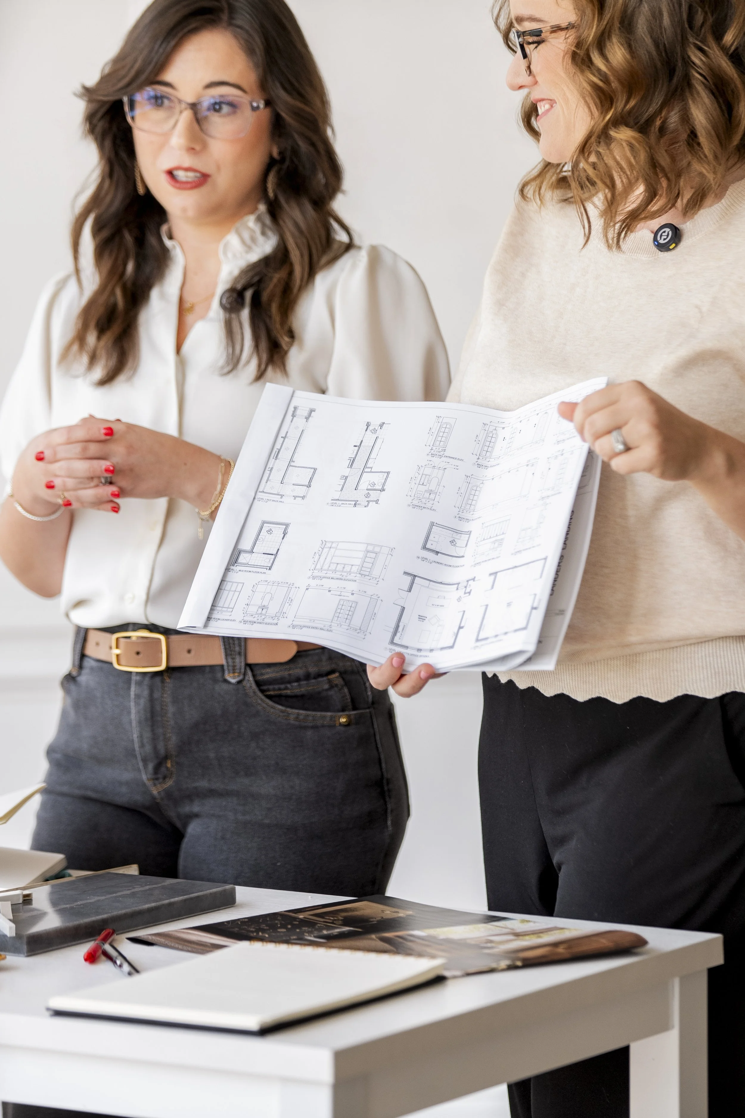Two women discussion and sharing architectural blueprints in an office, with a table displaying design materials and drawing tools.