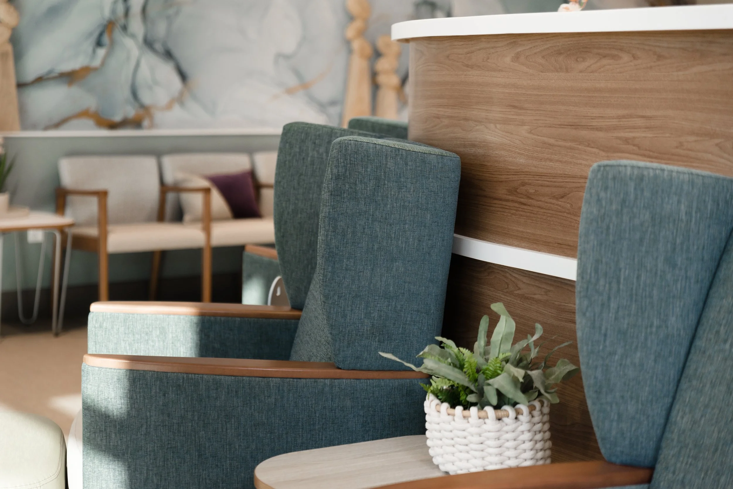 Close-up of a seating area with teal upholstered chairs and a wooden counter, a white wicker basket with a green plant inside, a modern decor background with a marble wall, and some wooden accents.