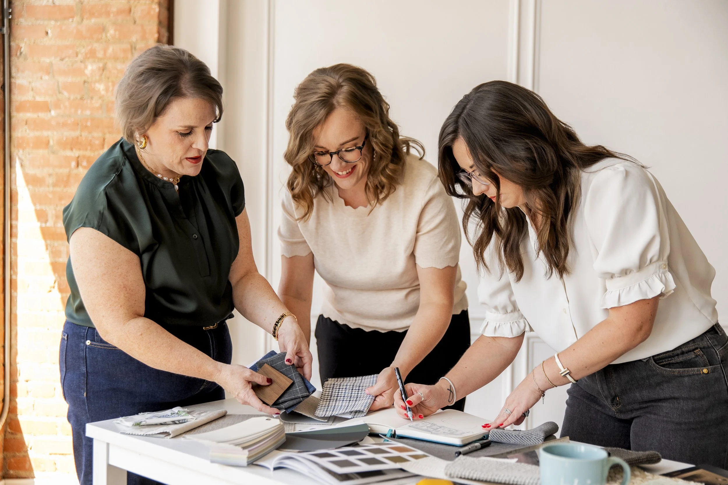 Three women collaborating on a project at a table covered with fabric swatches, color samples, and notebooks in a modern workspace.