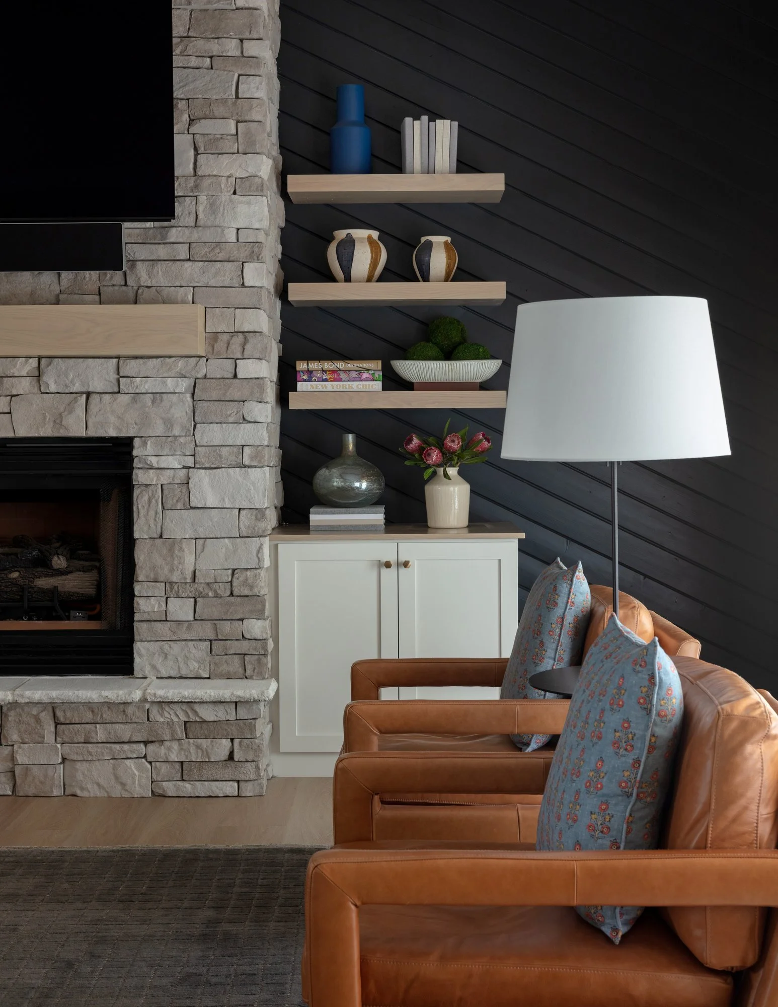 Living room with stone fireplace, black accent wall with floating shelves holding decorative vases and books, white cabinet with plant and decorative objects, leather chair with patterned pillows, floor lamp with large white shade, dark rug, and wooden flooring.
