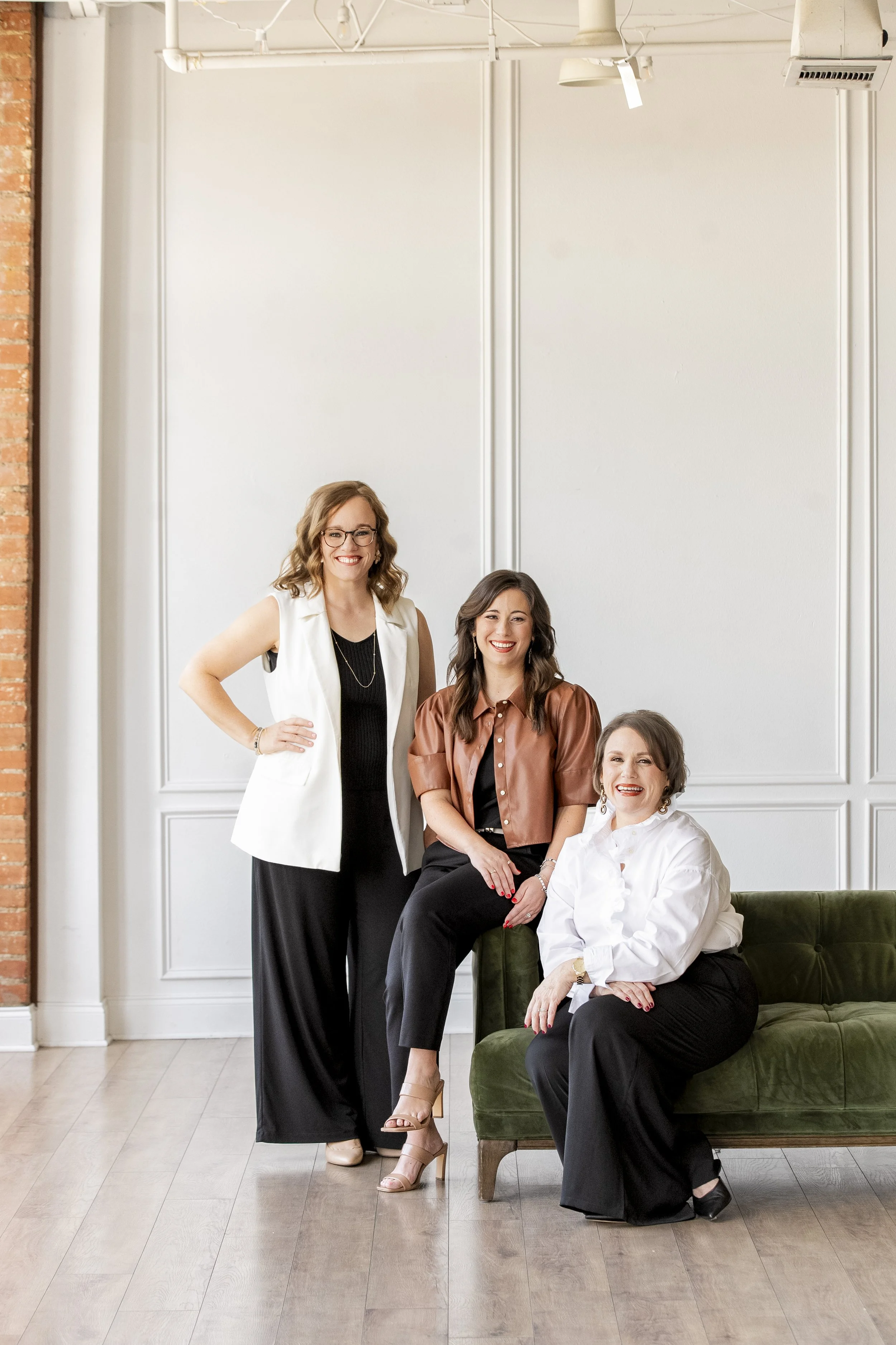 Four women in business attire posing in a modern, minimalist room with white walls and wooden floors. One woman is standing while the others are sitting on a green velvet sofa.