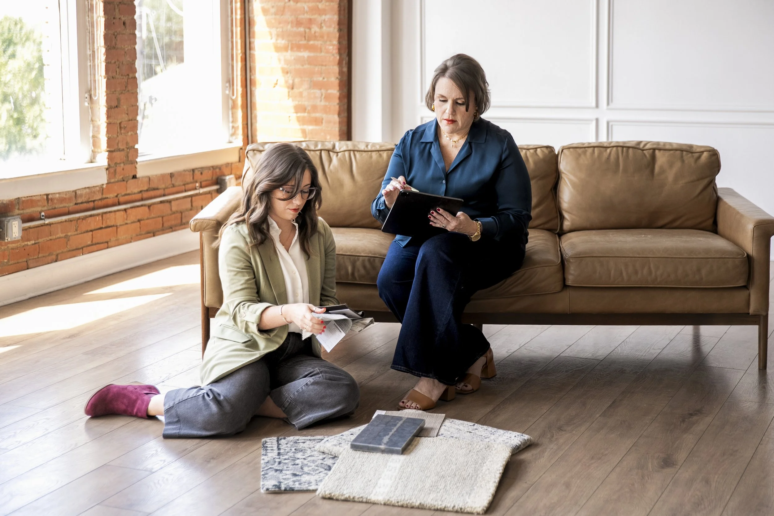 Two women in a bright room with large windows, one sitting on the floor with reading glasses and papers, the other sitting on a couch, looking at a tablet.