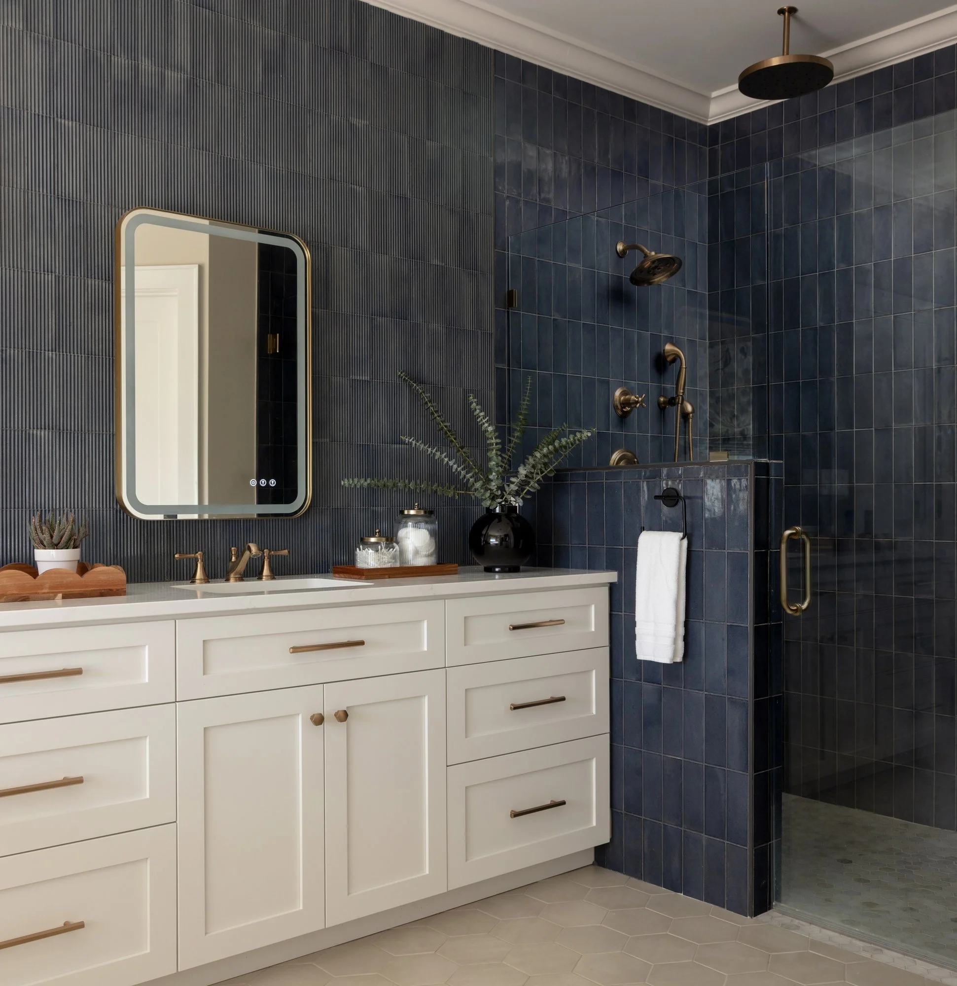 Modern bathroom with a white vanity featuring gold handles, a black vase with greenery, a small potted succulent, and a soap dispenser, against a dark blue tiled wall and a walk-in shower with black fixtures and a gold rain showerhead.
