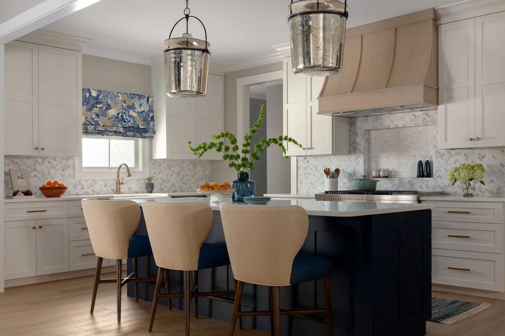 A modern kitchen with white cabinetry, a black island counter, beige upholstered chairs, and hanging metallic pendant lights. There is a window with a blue patterned valance, decorative plants, and kitchen utensils on the countertop.