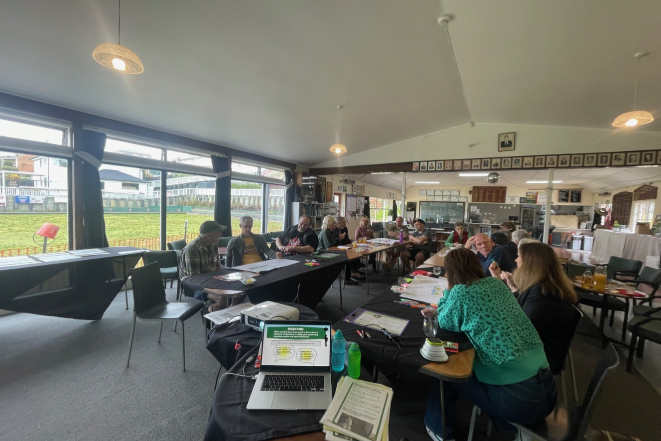 A hall full of people sits in a u-shaped formation of tables, all discussing and working together on some mahi. There is a laptop with a presentation on it on a small table at the front, and another table to the left back of the hall with paper on it