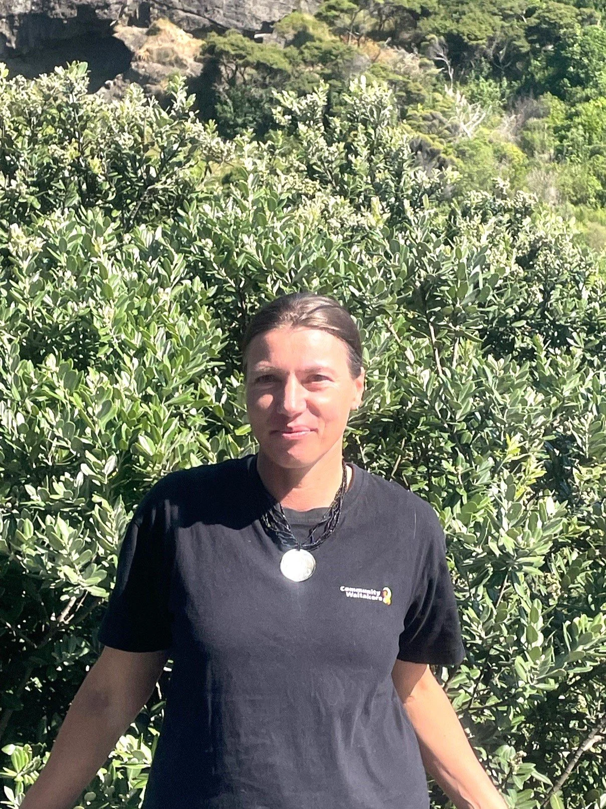 Shalema Wanden-Hannay, a woman standing outdoors in front of large green shrub, wearing black t-shirt and necklace, with trees and a hillside in the background.