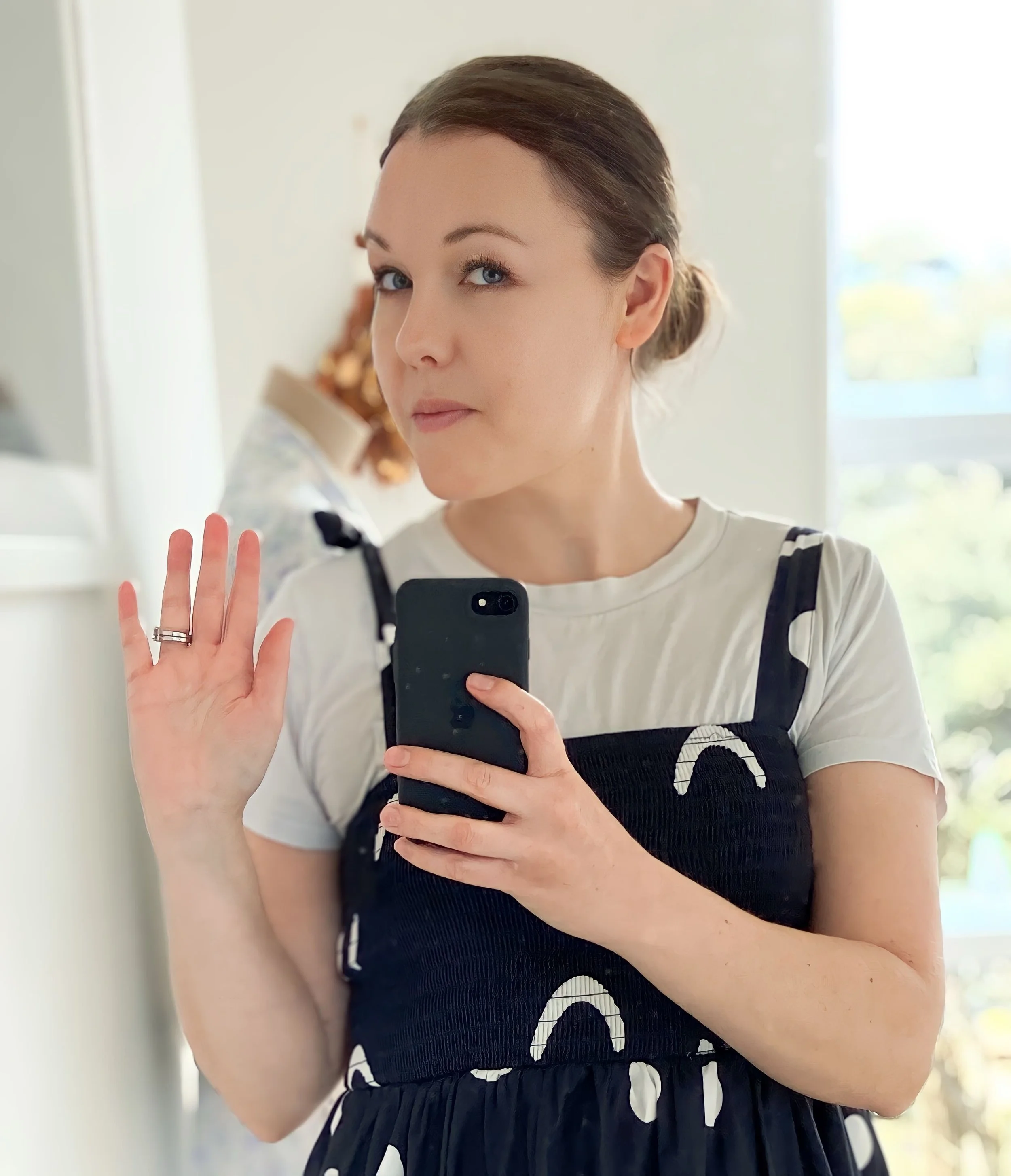 Sarah Mikkelsen, a woman with light skin and brown hair in a bun, taking a mirror selfie whilst waving with one hand and holding her phone in the other.