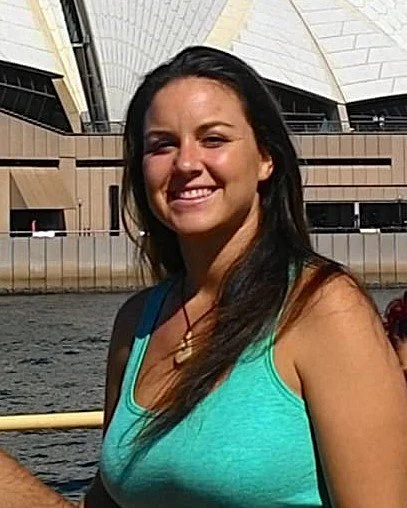 Anna Berthiaume, a woman with long dark hair smiling outdoors near water, with the Sydney Opera House in the background.