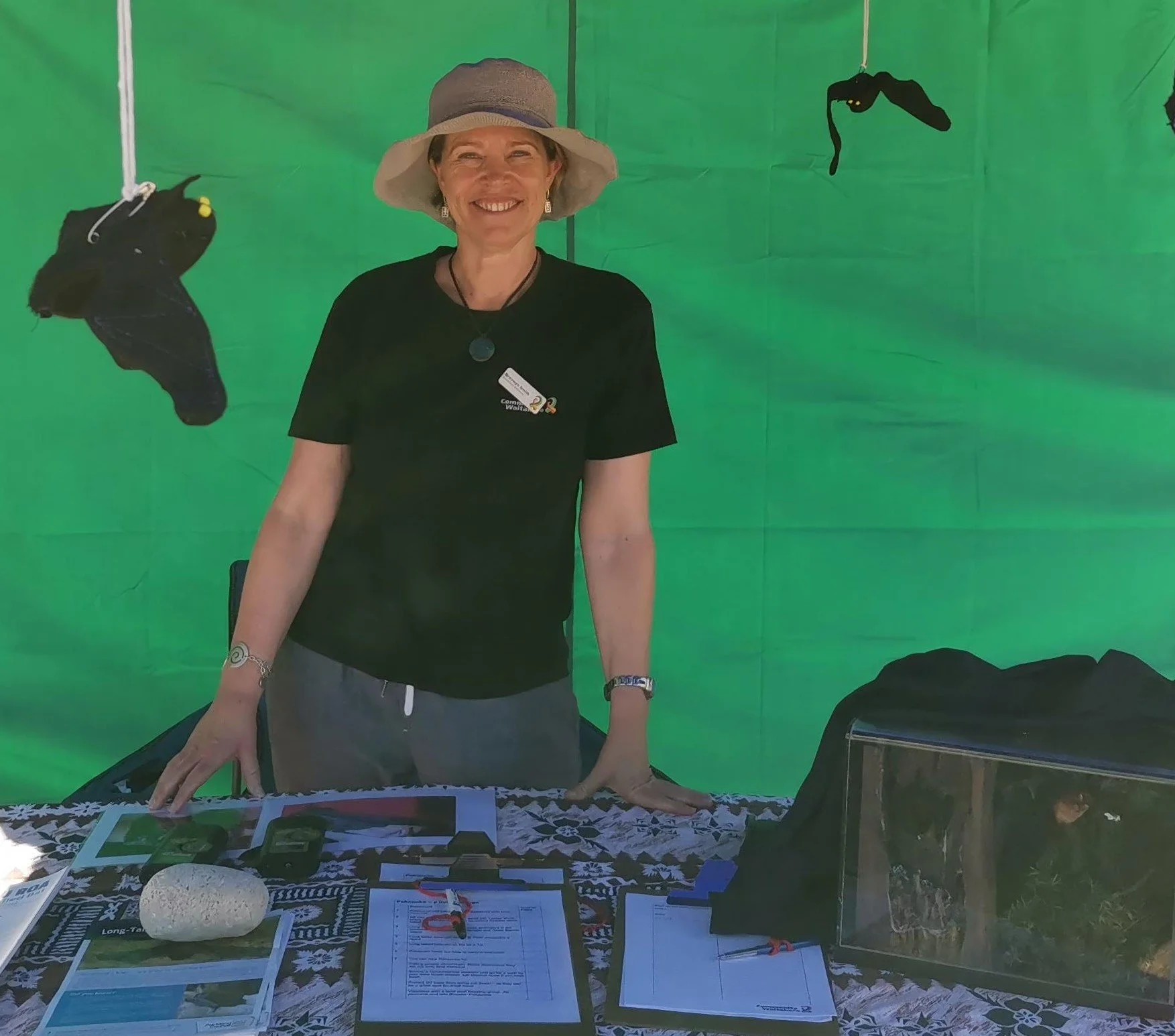 Bronwyn Smith, a woman standing behind an information booth with a green backdrop and black bats hanging above her, wearing a black shirt, a hat, and jewelry, smiling at the camera.