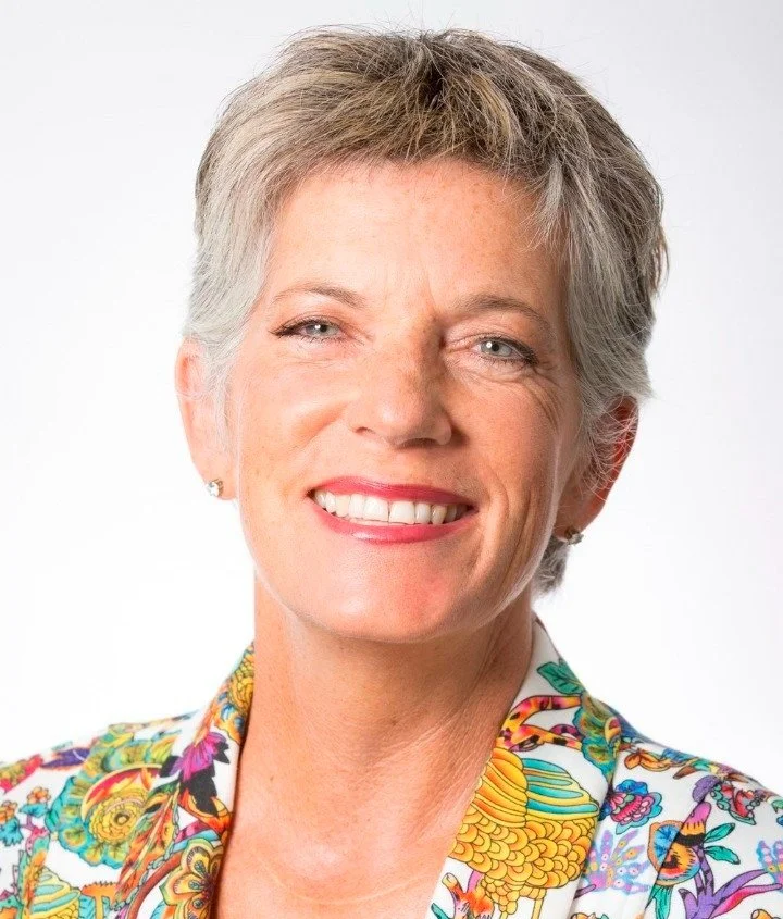 Penny Hulse, a middle-aged woman with short gray hair smiling, wearing a colorful, patterned blazer against a plain white background.