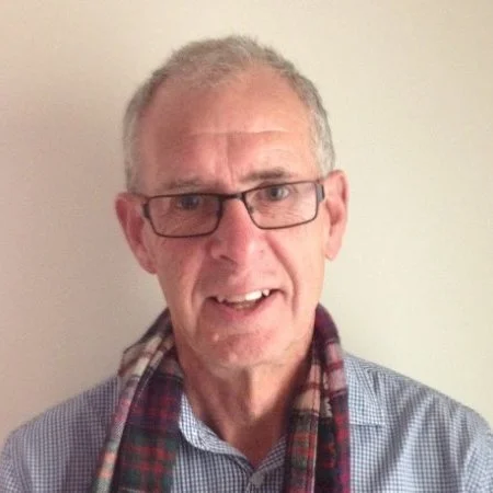 Charlie Moore, a middle-aged man with gray hair and glasses, wearing a checkered shirt and a plaid scarf, smiling in front of a plain light-colored wall.