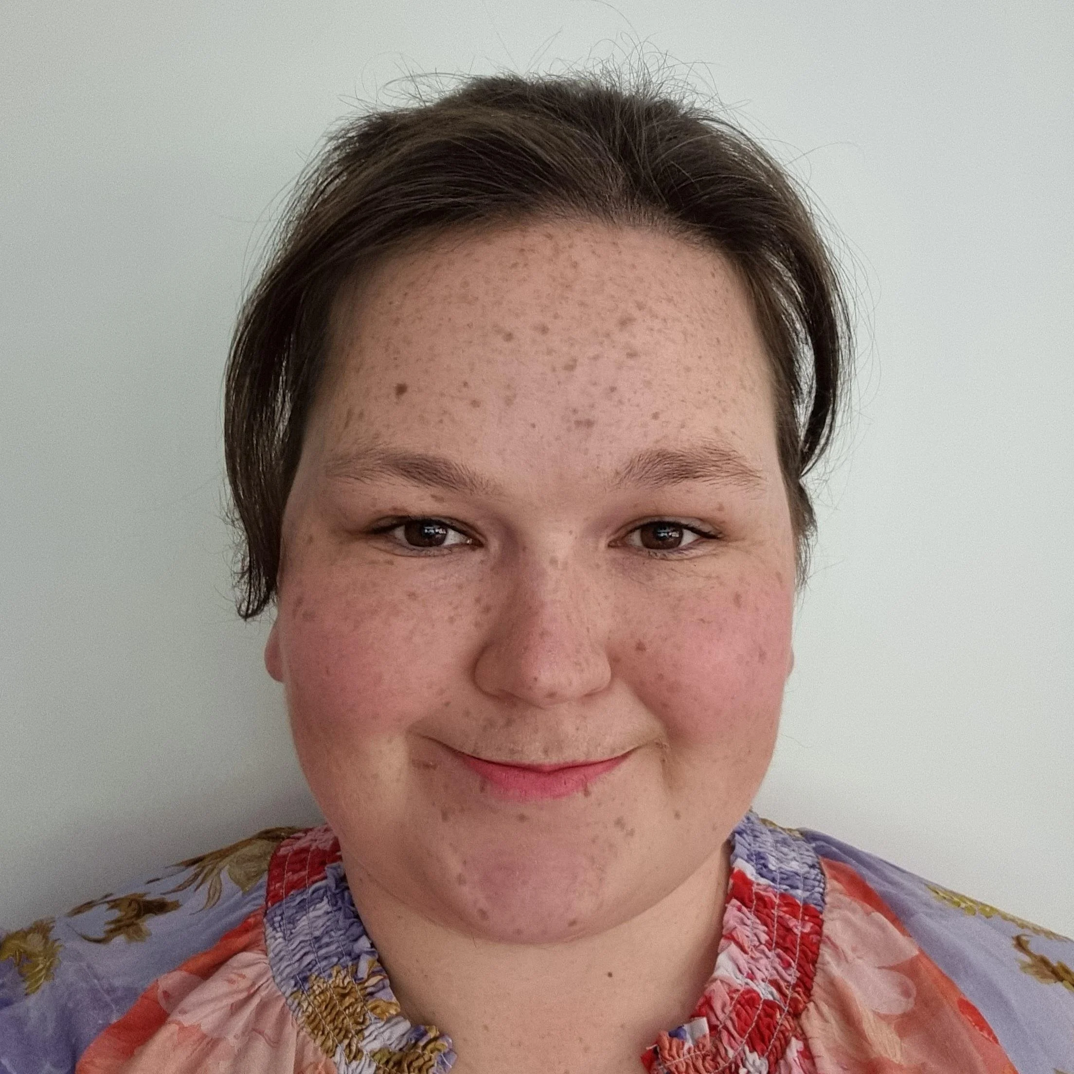 Close-up of Chelsea Oswin, a smiling woman with short brown hair, freckles, and wearing a floral top, standing against a plain white wall.