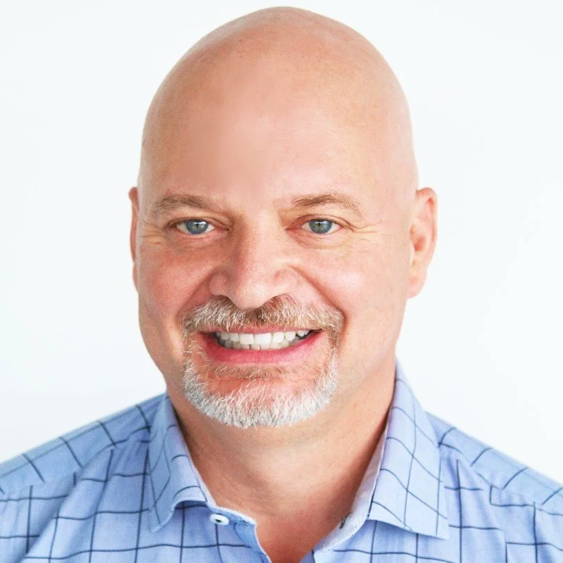 Headshot of Peter Young, a middle-aged man with a bald head, blue eyes, and a trimmed white beard, smiling and wearing a light blue checkered shirt.