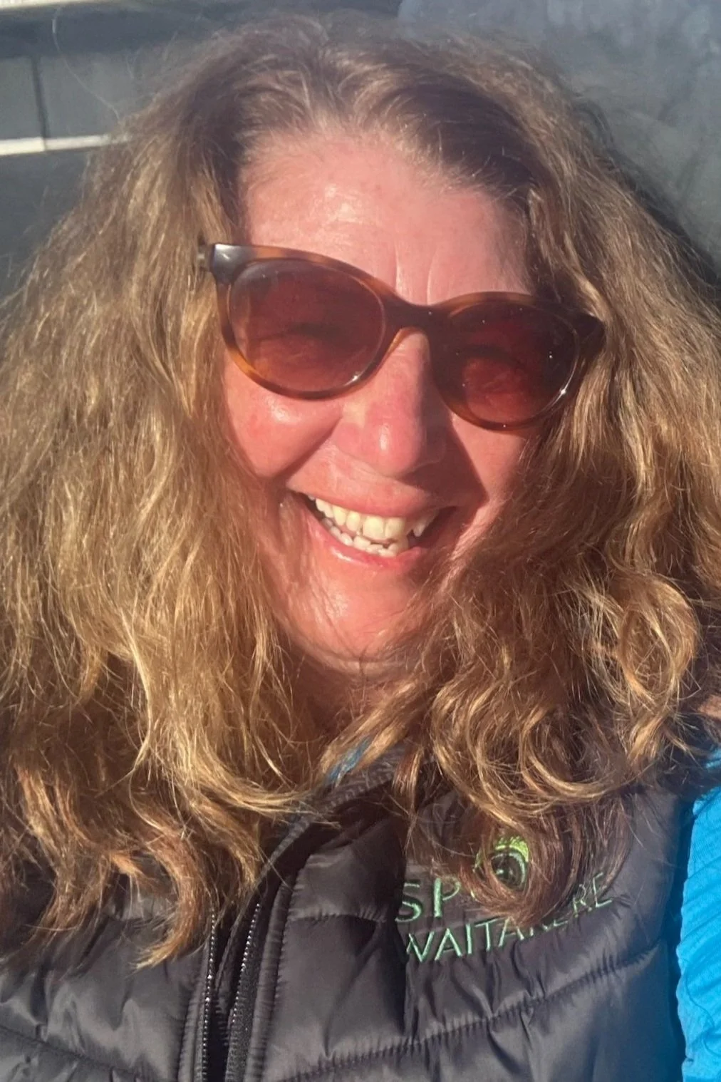 Kerry Allan, a woman with shoulder-length curly brown hair standing against a teal background, wearing a patterned blouse with long, puffy sleeves, a dark skirt, a watch on her left wrist, and a ring on her right hand.