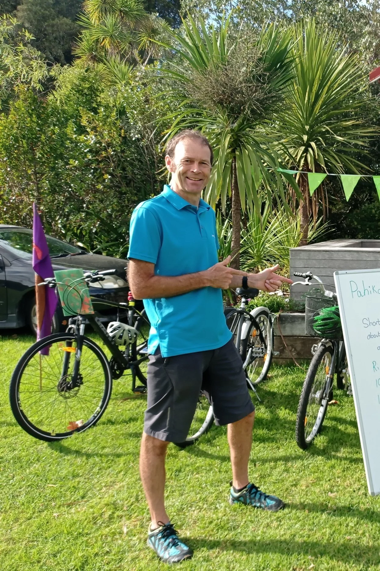 Simon Vincent, a man in a blue shirt and shorts pointing at a whiteboard outdoors with bicycles and lush greenery in the background.