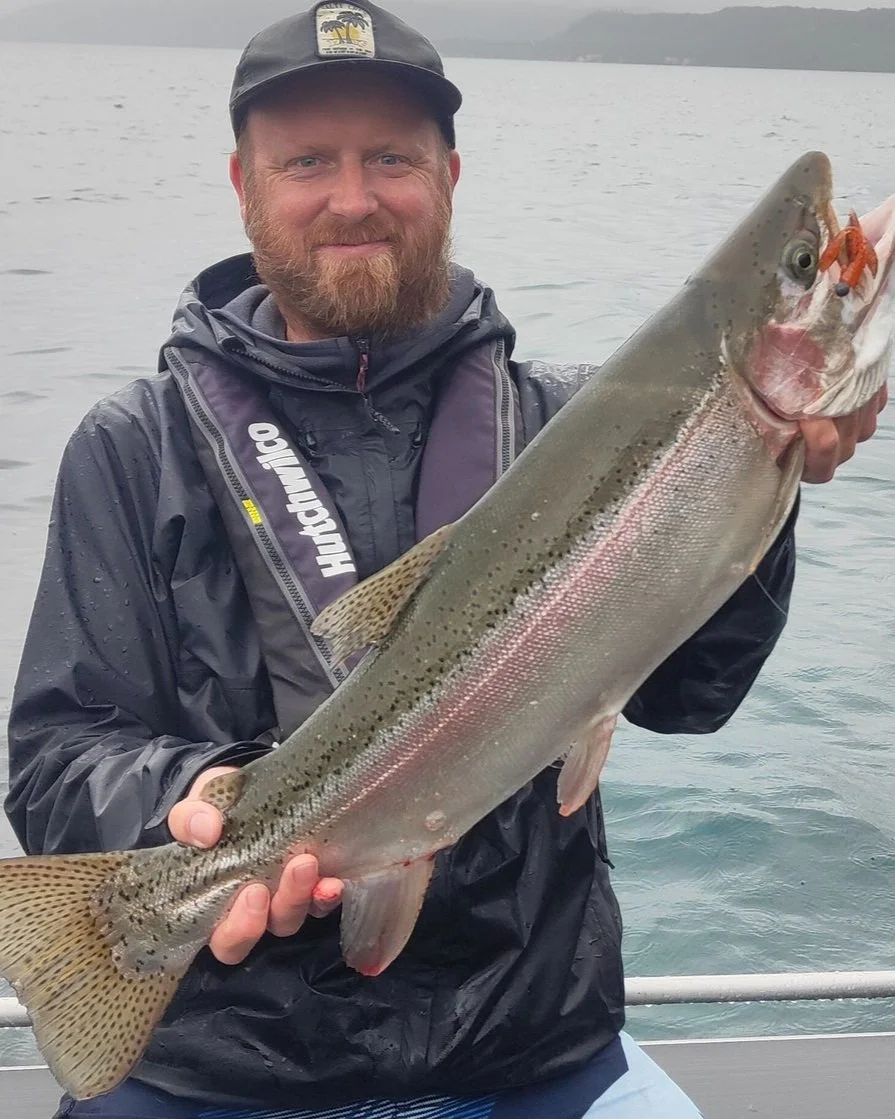 Chris Burton, holding a large rainbow trout on a boat in a lake, wearing a Cap and waterproof jacket.