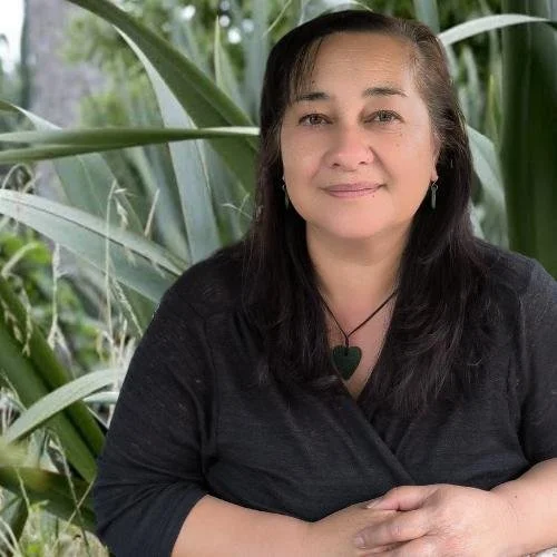 Daena Walker, a woman with long dark hair wearing a black shirt and a green heart-shaped necklace, sitting outdoors among harekeke (NZ flax).