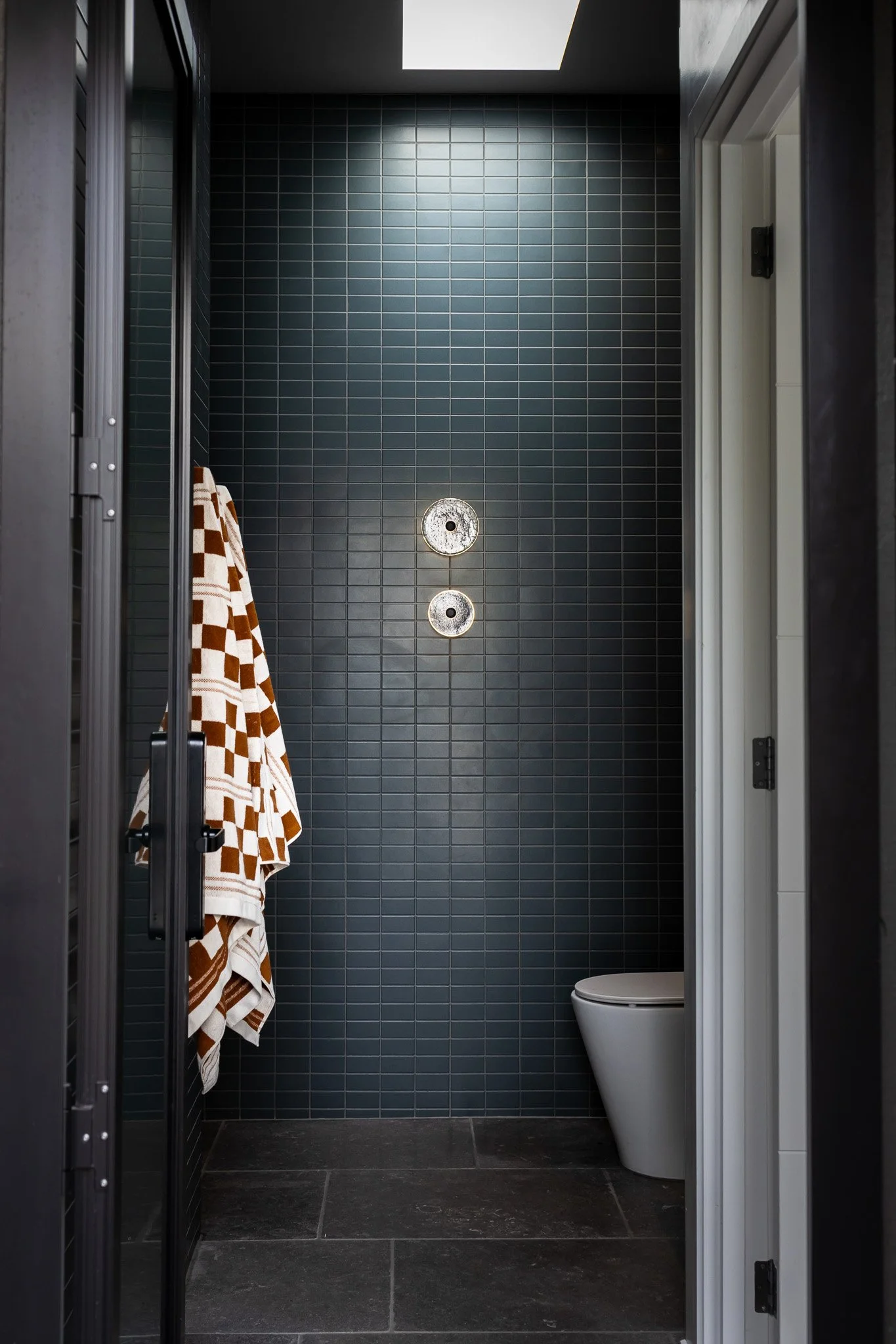 Empty modern shower with dark gray tiles, a towel hanging on a hook, and a partly visible white toilet in a bathroom.