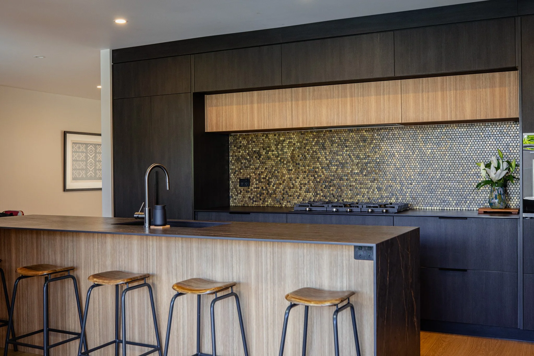 Modern kitchen with black cabinets, a wooden island with four bar stools, a sink, a gold mosaic backsplash, a gas stove, and a vase with white flowers.